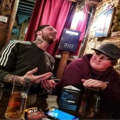 Two men sitting at a wooden table in a cozy bar, laughing and talking. One has tattoos and is wearing a black Adidas jacket, the other is wearing a baseball cap and a maroon hoodie. There are drinks, a smartphone, and some objects on the table. The b
