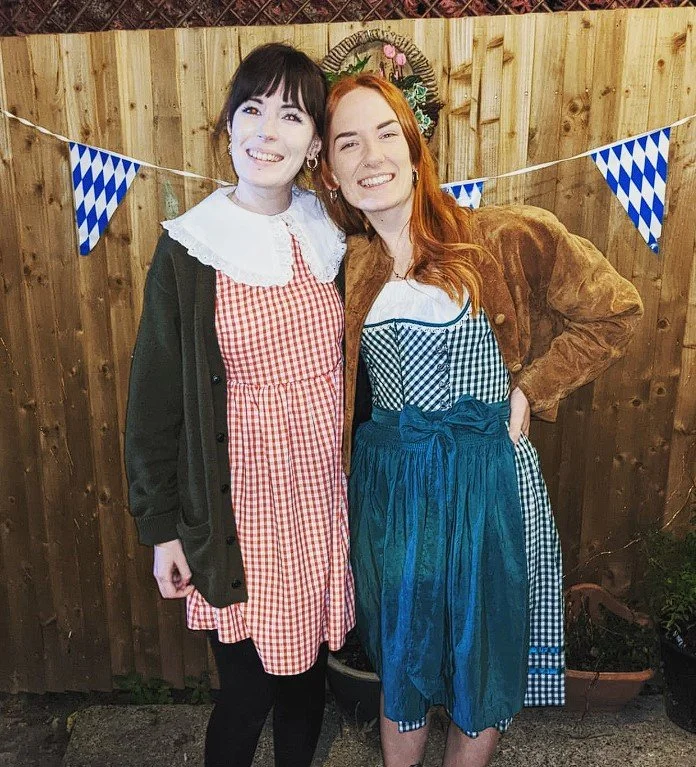 Two women standing in front of a wooden fence decorated with blue and white checkered bunting. One woman has dark hair and is wearing a red and white checkered dress with a white doily collar, and a black cardigan. The other woman has red hair and is wearing a blue plaid dress with a blue apron and a brown jacket.