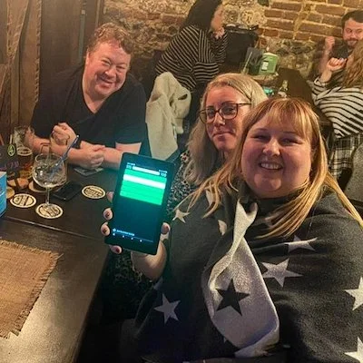 Three people sitting at a bar, with two women in the foreground, one holding a smartphone showing a green screen, and a man smiling in the background, along with other bar patrons and a brick wall.