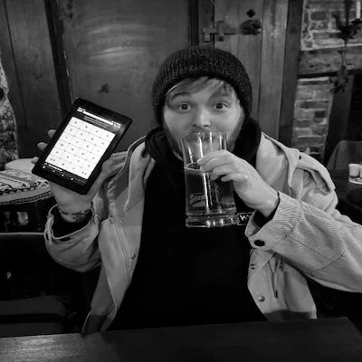 Person in a beanie and jacket holding a smartphone in one hand and a glass of soda in the other, sitting at a wooden table in a rustic setting