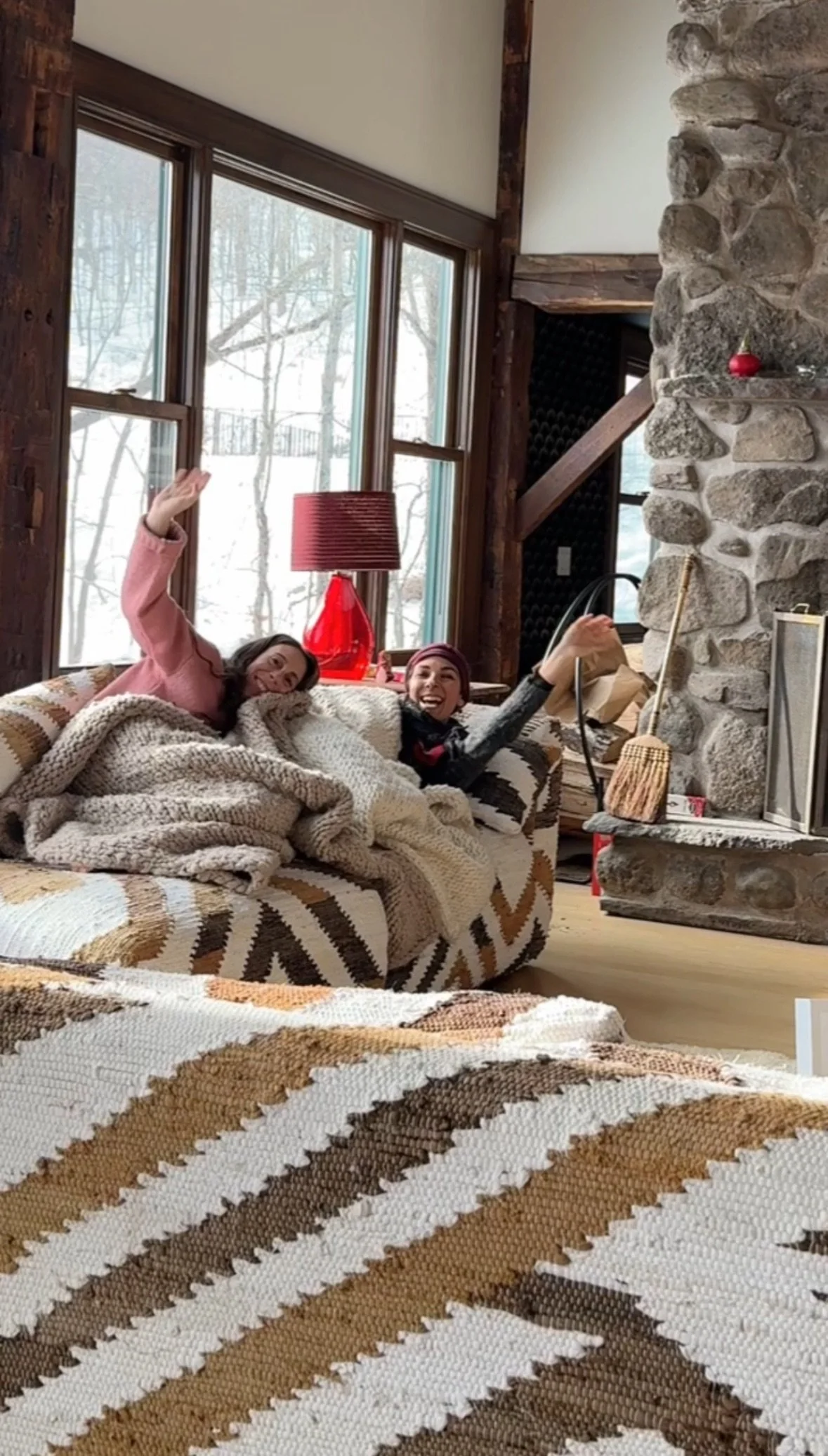 Two women sitting on a patterned sofa in a cozy living room, smiling and waving, with snow visible outside the large windows behind them, a red lamp on the side table, and a stone fireplace with a broom resting against it.