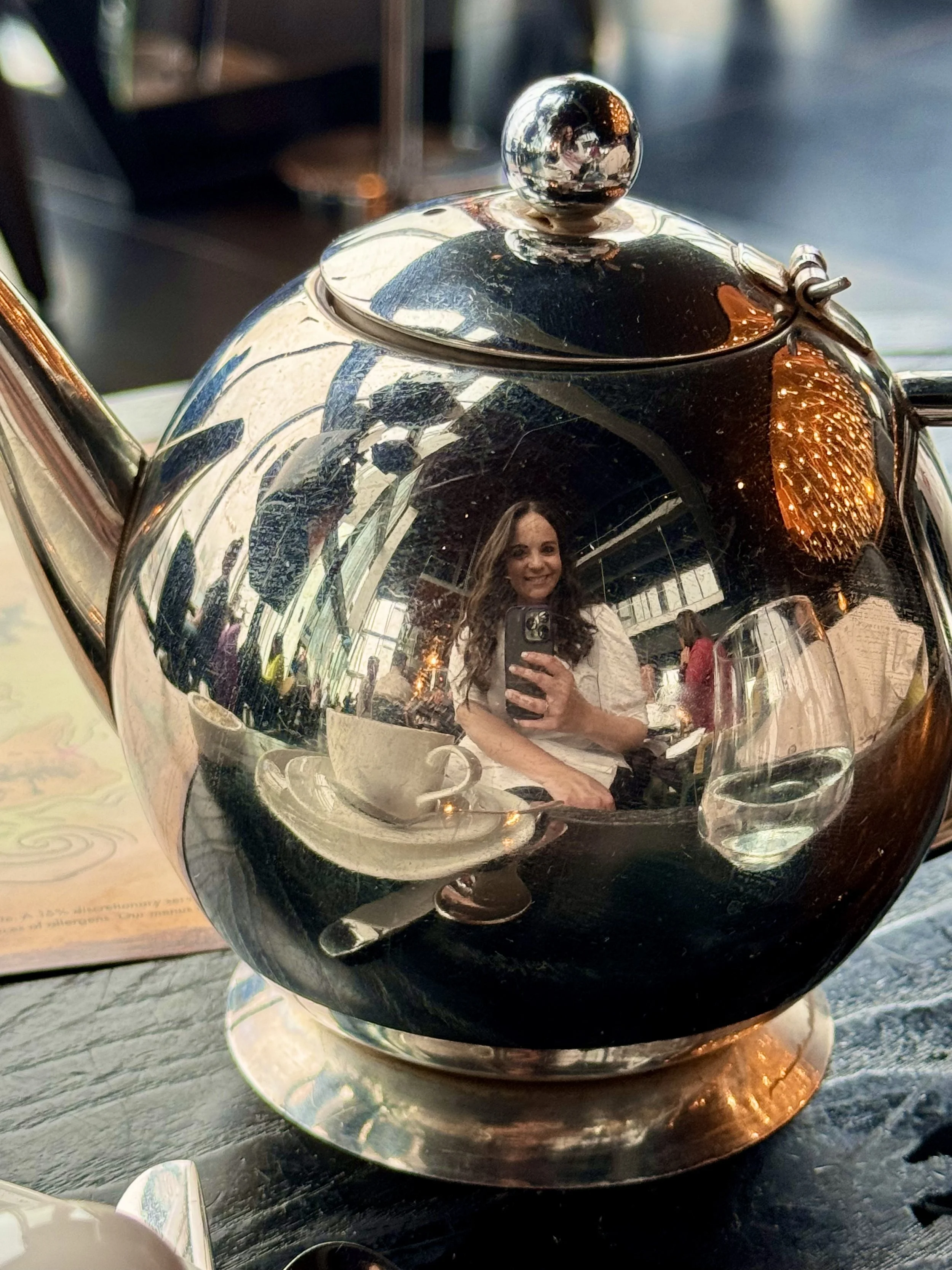 A smiling woman takes a selfie with her cell phone in the reflection of her tea pot