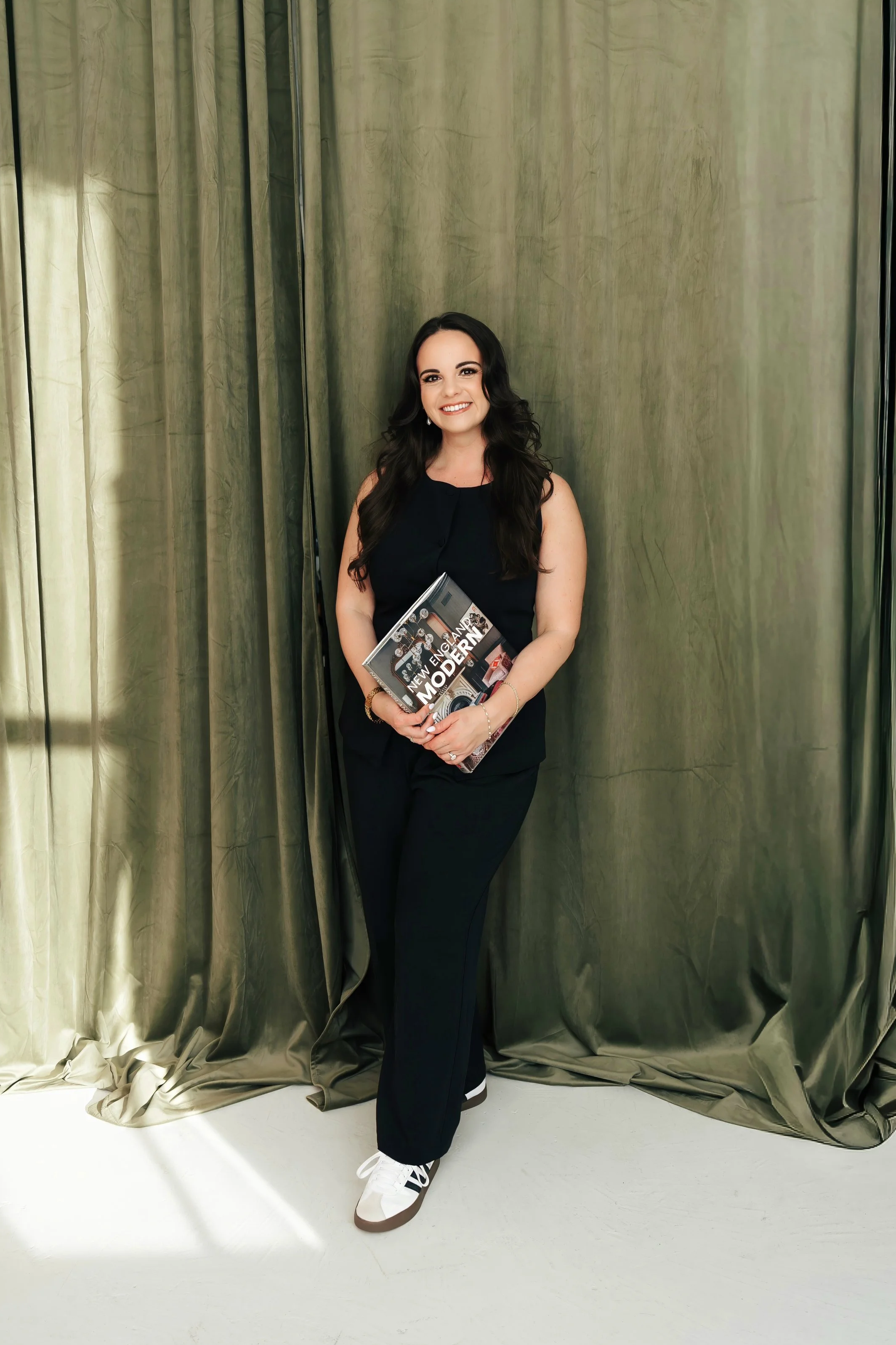 Smiling woman in black jumpsuit holds a design book in front of green curtains