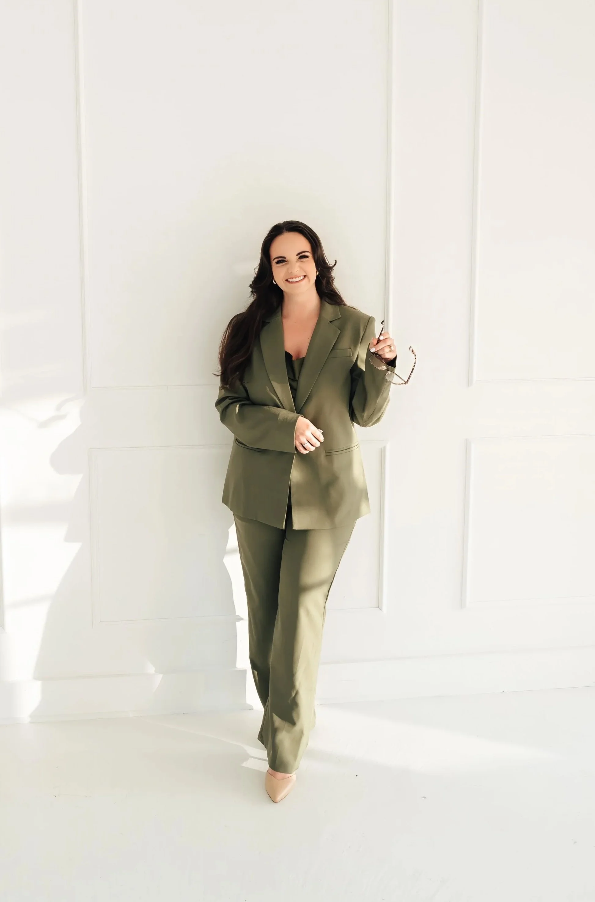 Woman in olive suit leans against a white wall holding a pair of glasses