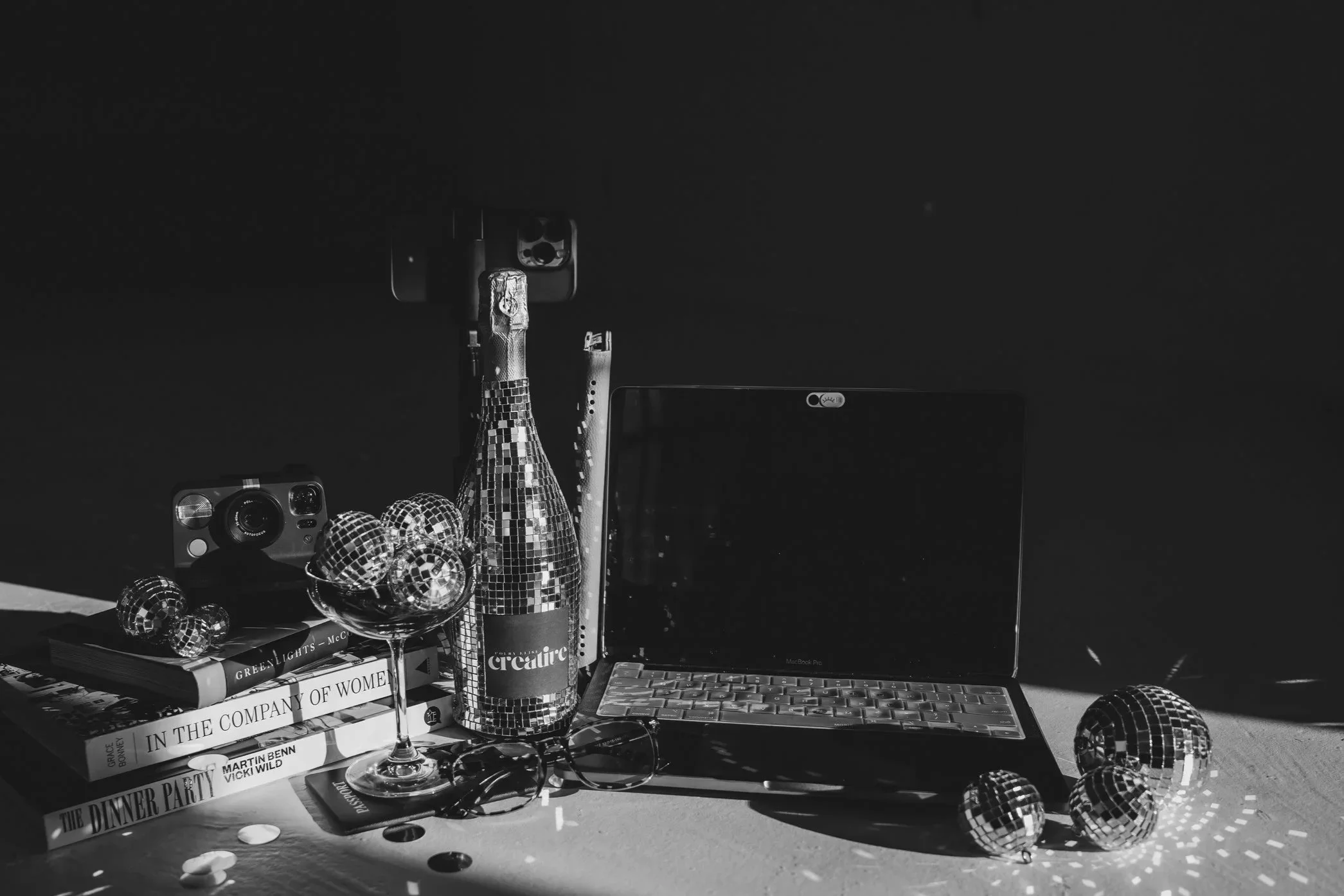 Black and white photo of a tablescape with a laptop, champagne bottle, disco balls, polaroid camera and books
