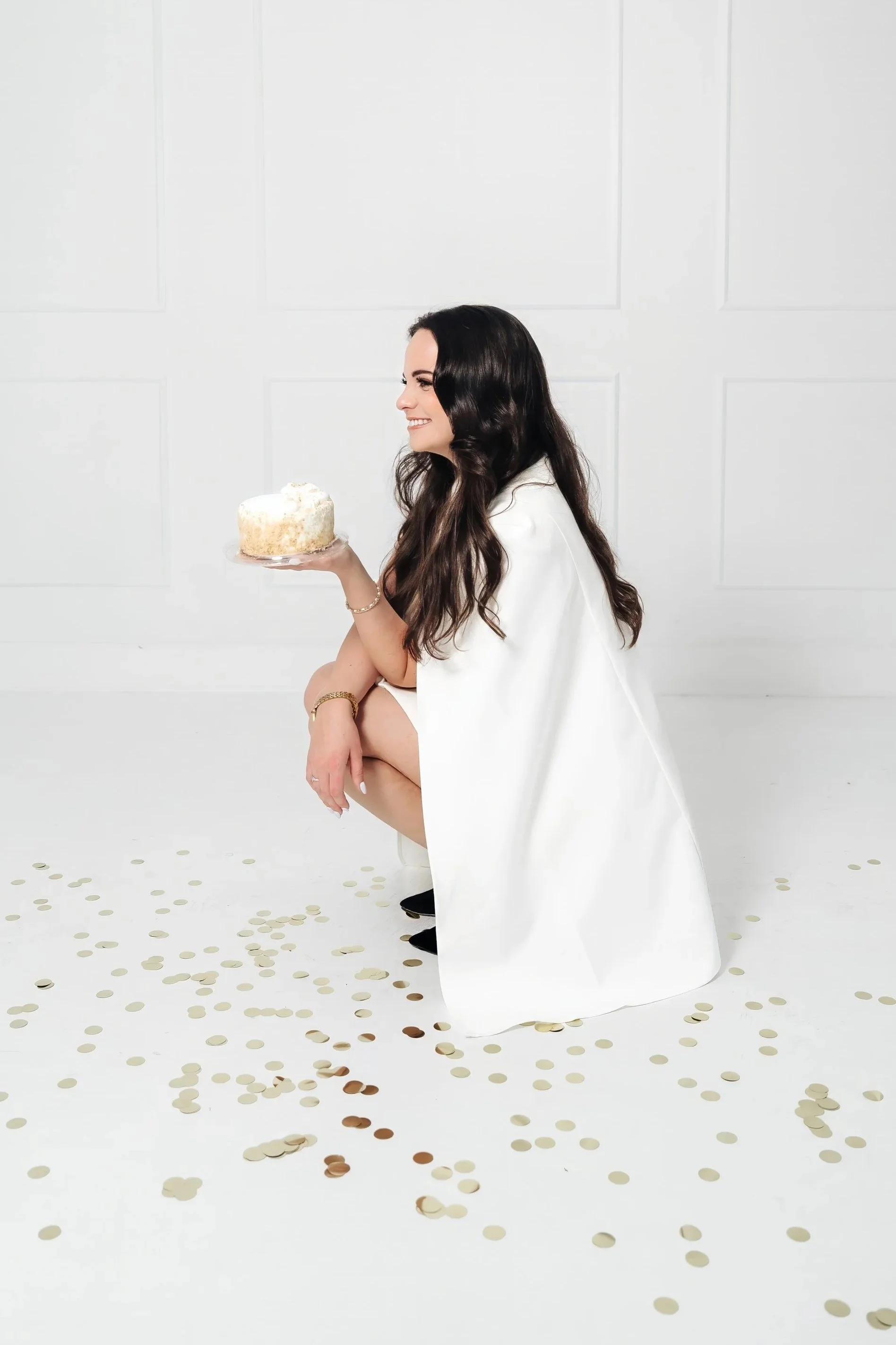 Smiling woman in white cape squats down holding a mini birthday cake