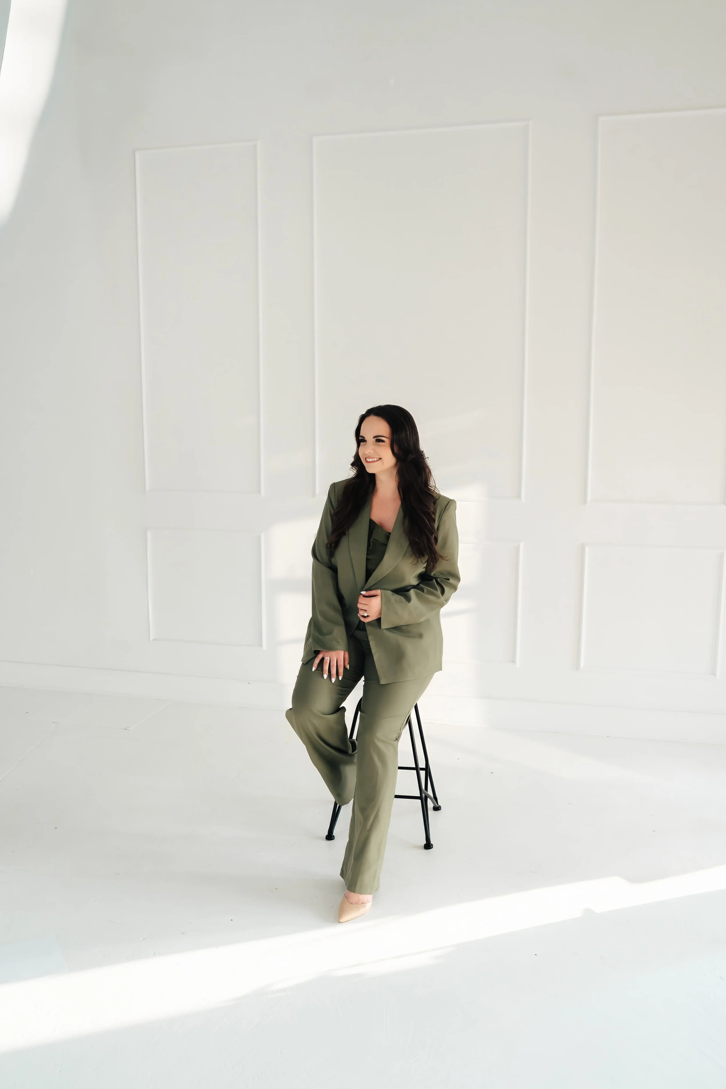 Smiling woman in a green suit sitting on a stool against a white wall