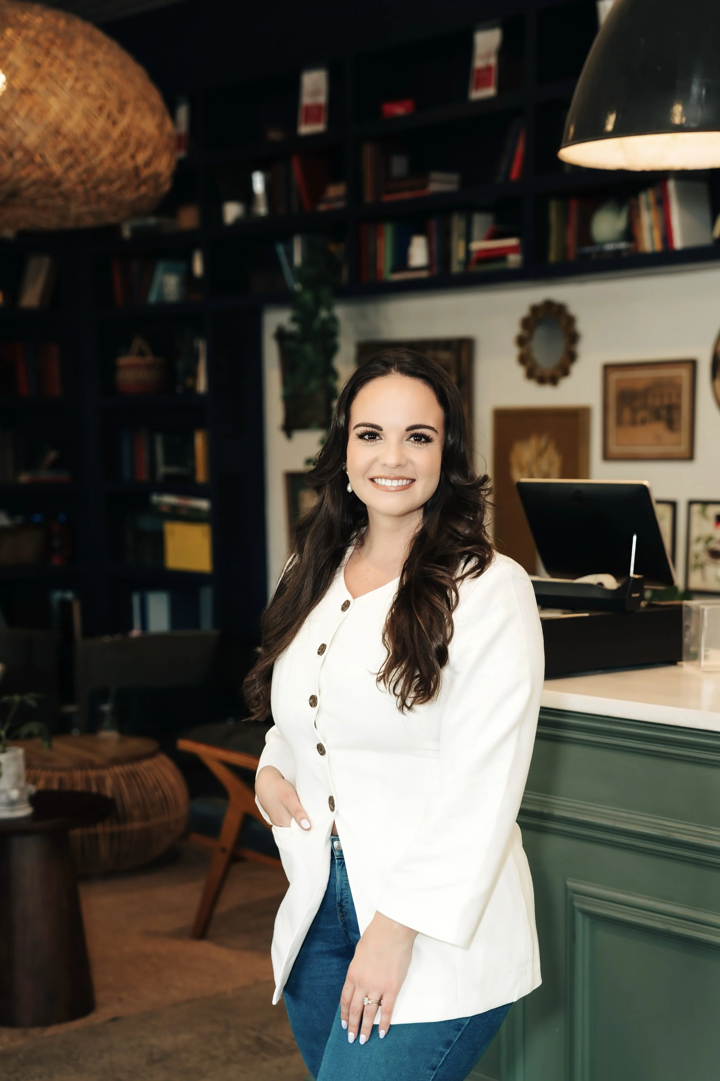 Smiling woman in white jacket and jeans stands in front of a counter with her hand in her pocket