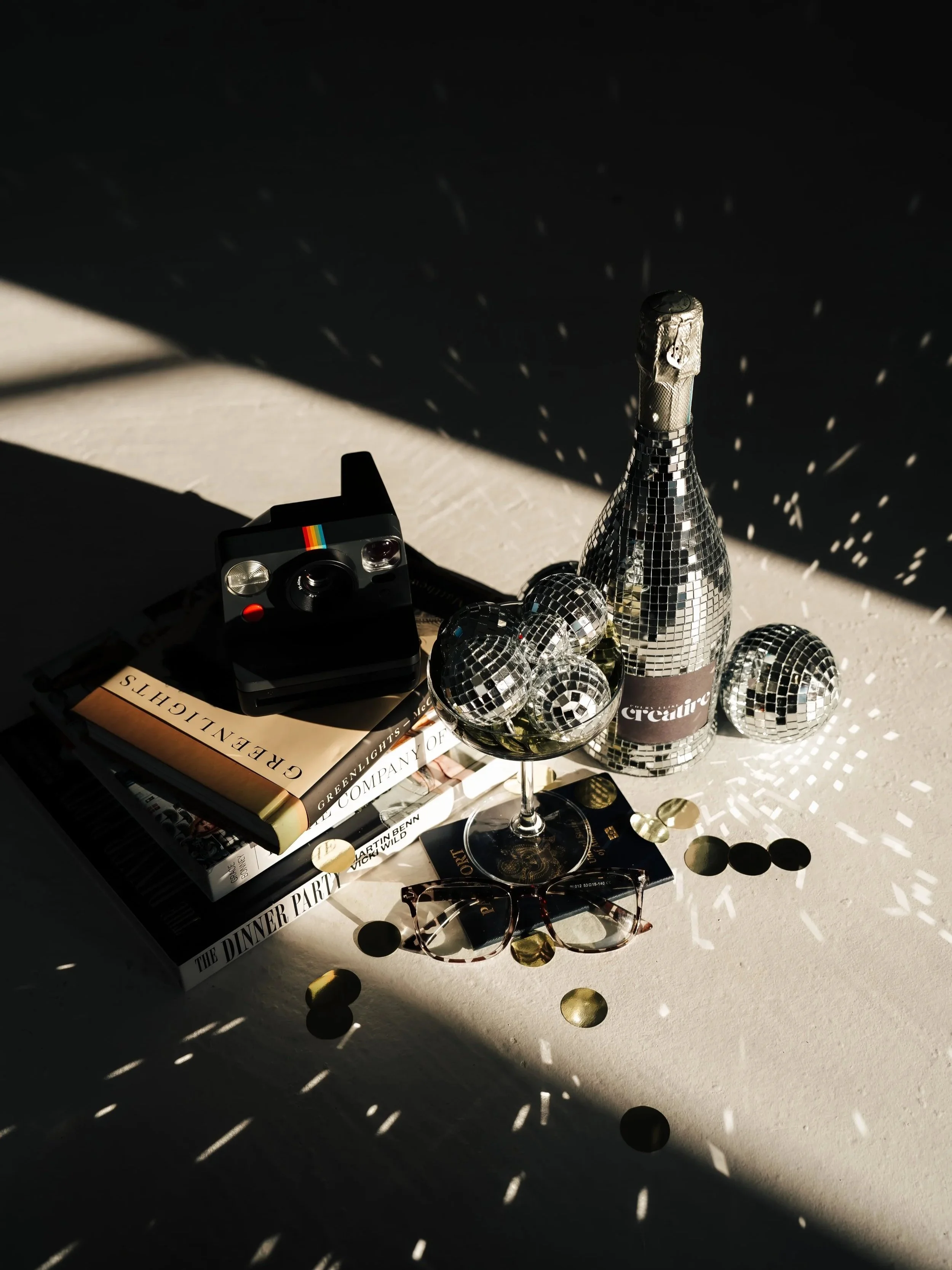 A polaroid camera rests on a stack of books next to disco balls in a glass and a disco ball champagne bottle