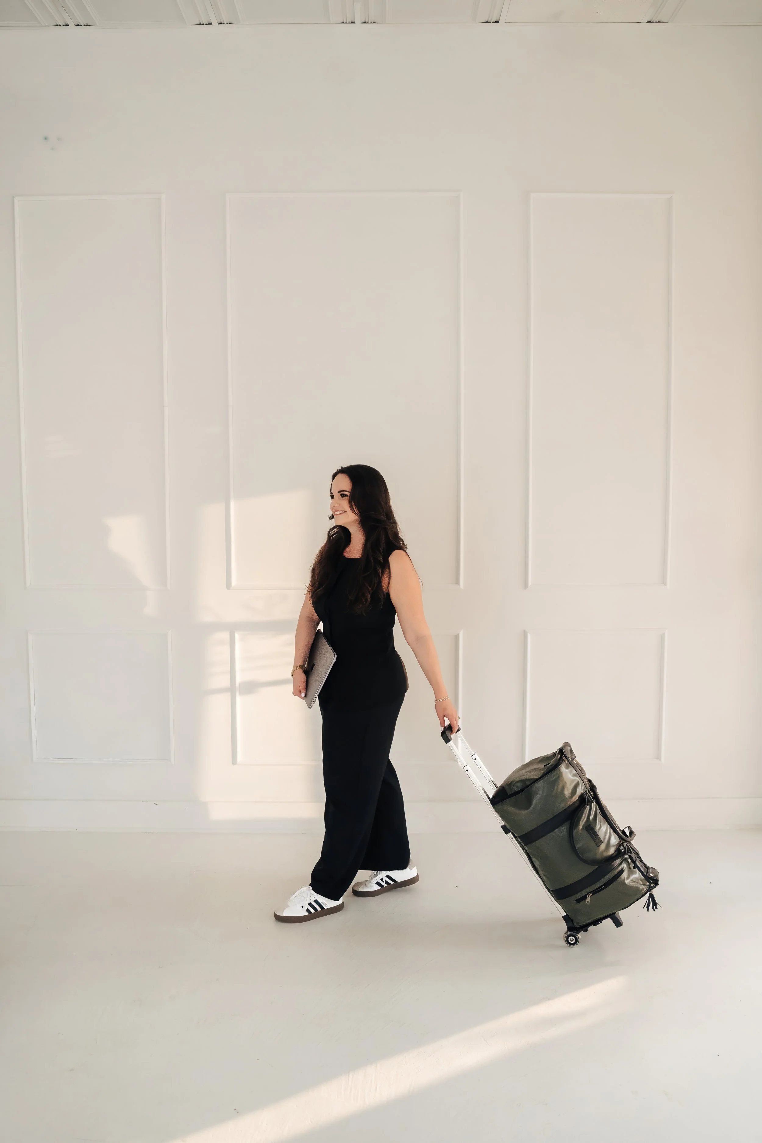 Woman in black jumpsuit holding a laptop and pulling a suitcase