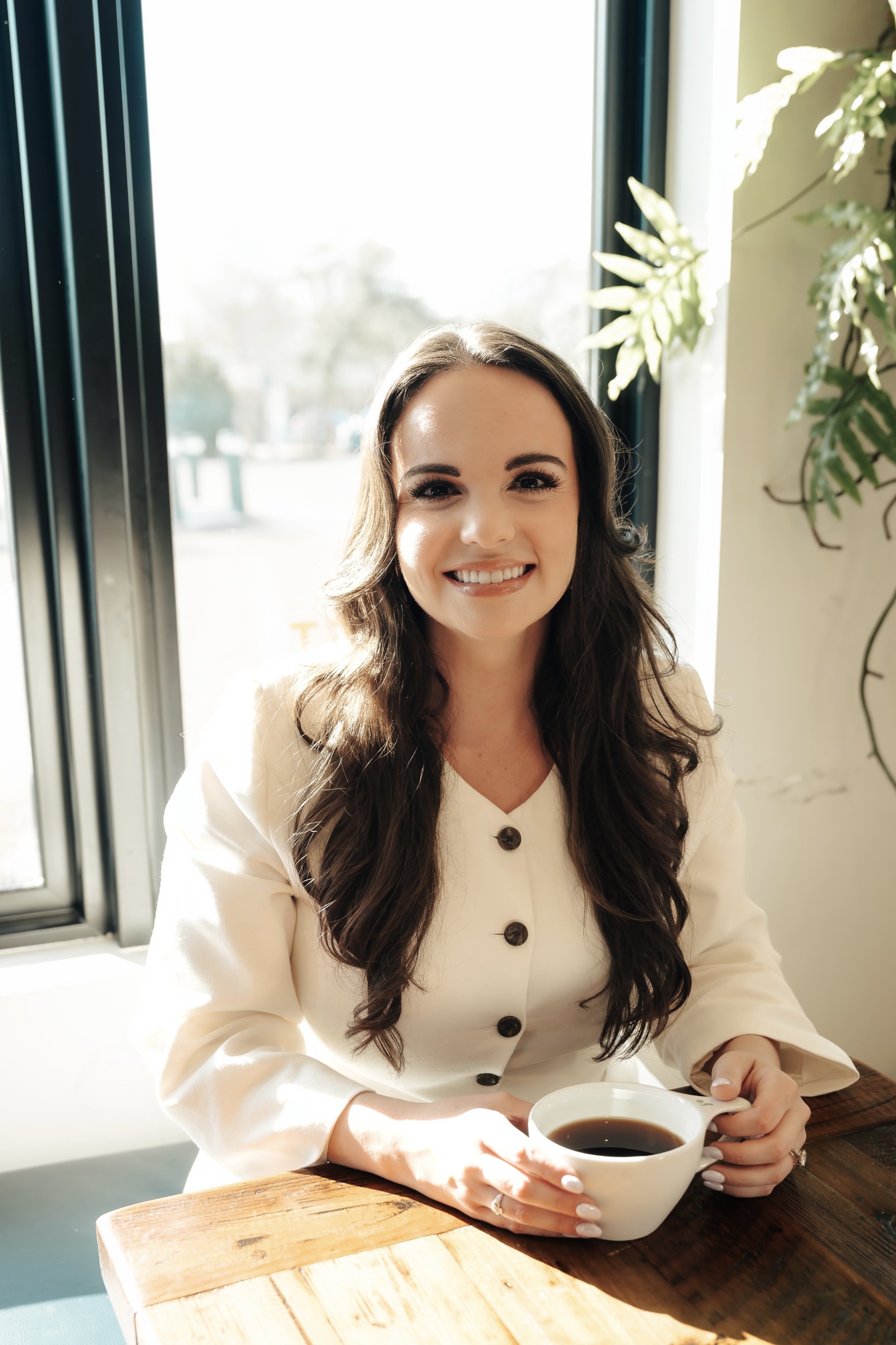 Woman in white jacket sits at a table in the window holding a coffee cup