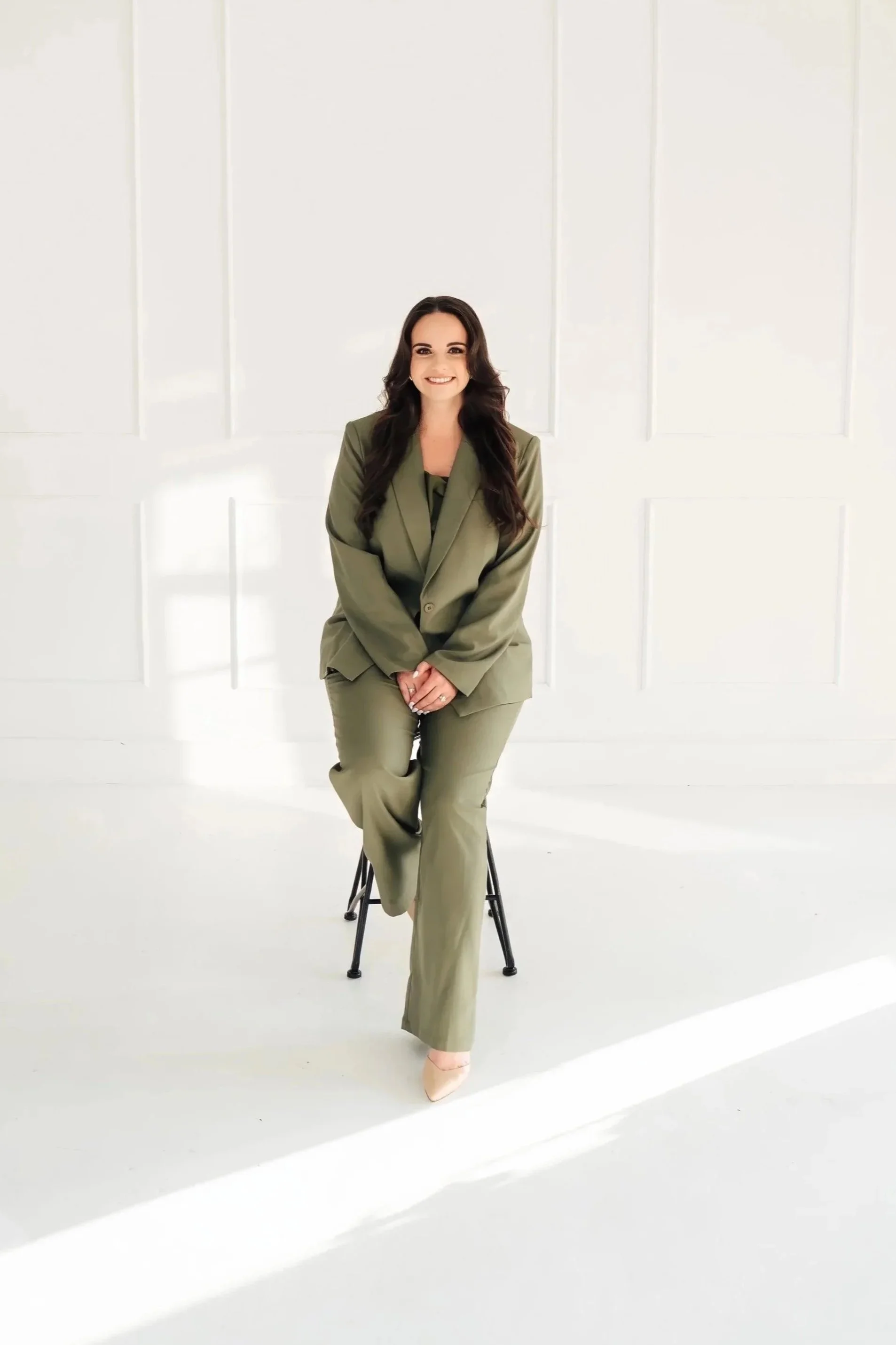 Woman wearing olive green suit smiles into the camera while sitting on a stool