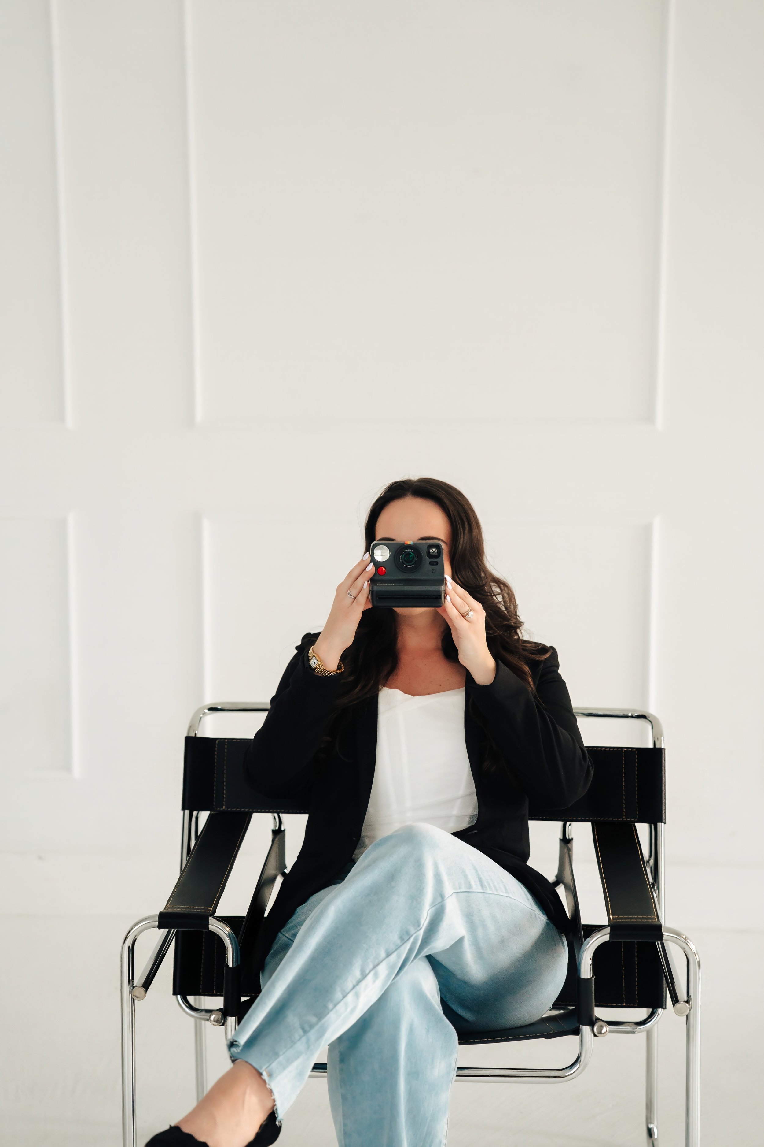 Woman sitting in chair in jeans and black blazer holding a polaroid camera