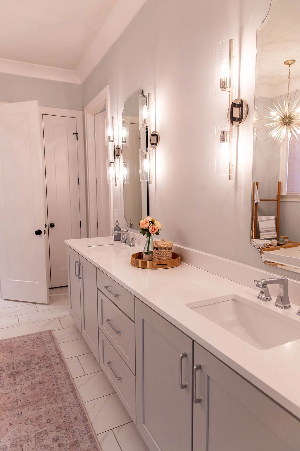 Bathroom with white double vanity, two sinks, large mirror, modern wall sconces, floral arrangement, wicker basket, light-colored tile floors, and a decorative rug.
