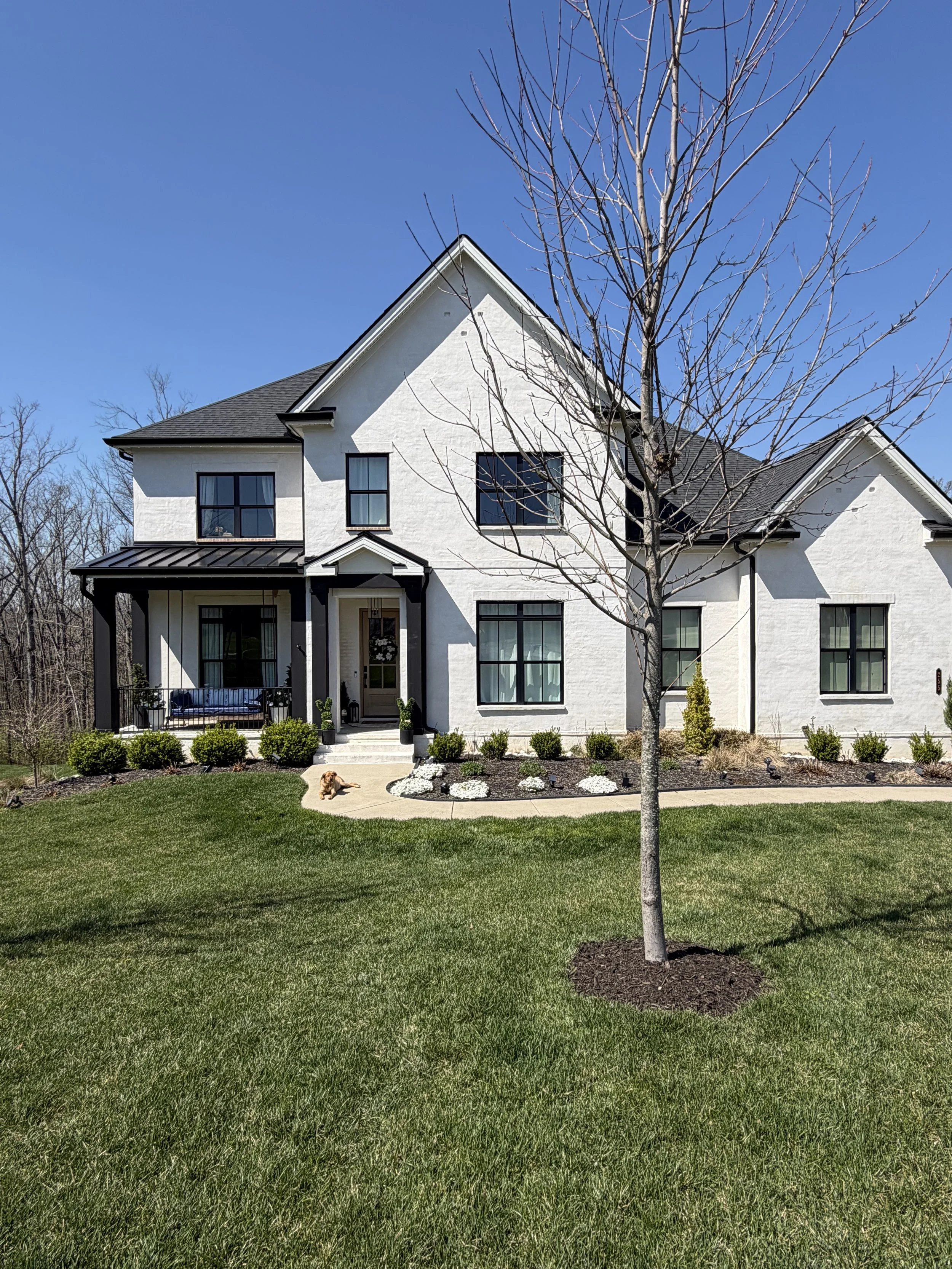 White two-story house with black window frames, front porch, and garden, in a yard with a green lawn, leafless tree, and a small dog near the pathway.