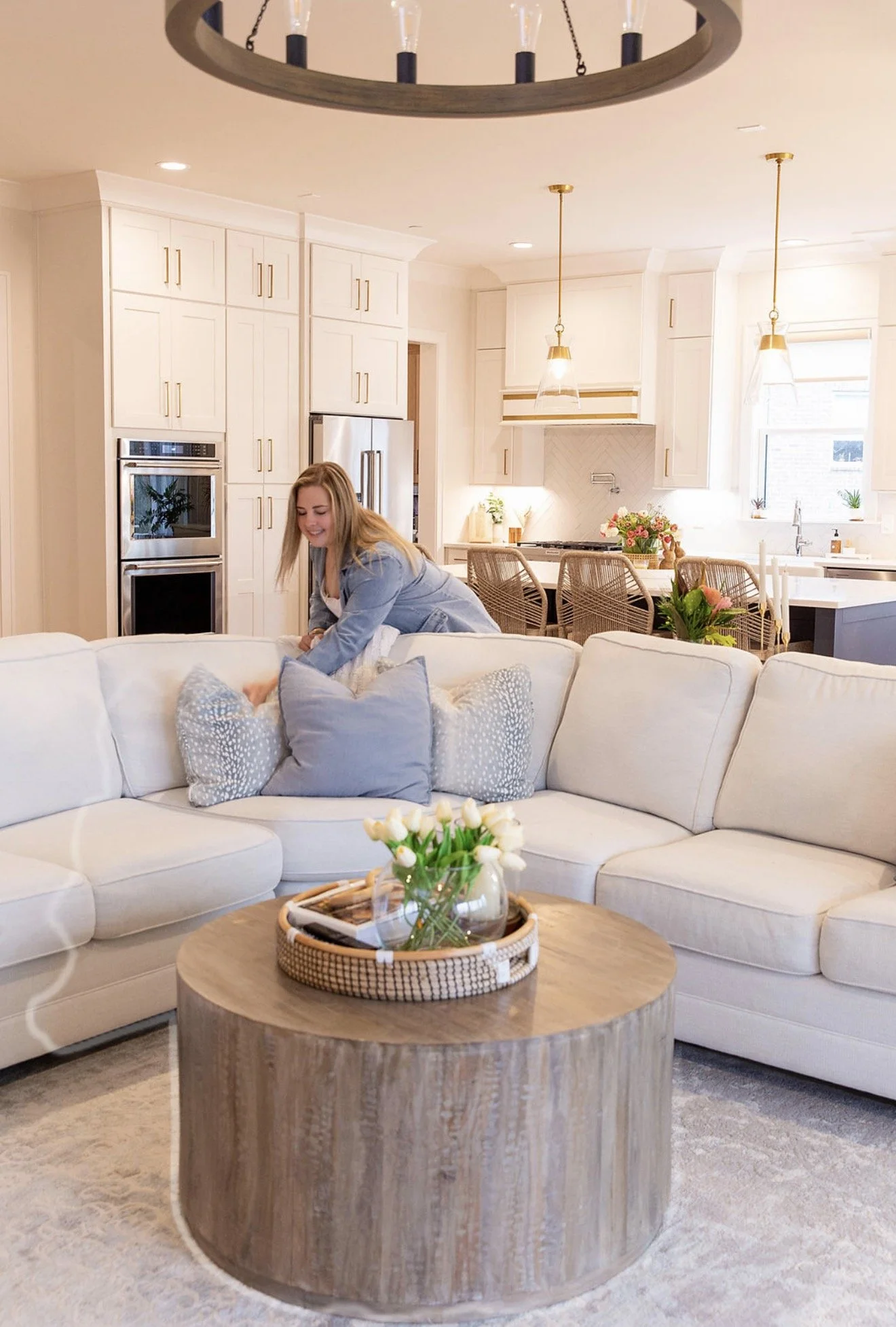 A woman smiling and leaning over a white sectional sofa in a bright, modern living room with a kitchen in the background. The room features white cabinets, a stainless steel refrigerator, a wooden coffee table with a vase of white tulips, and decorat