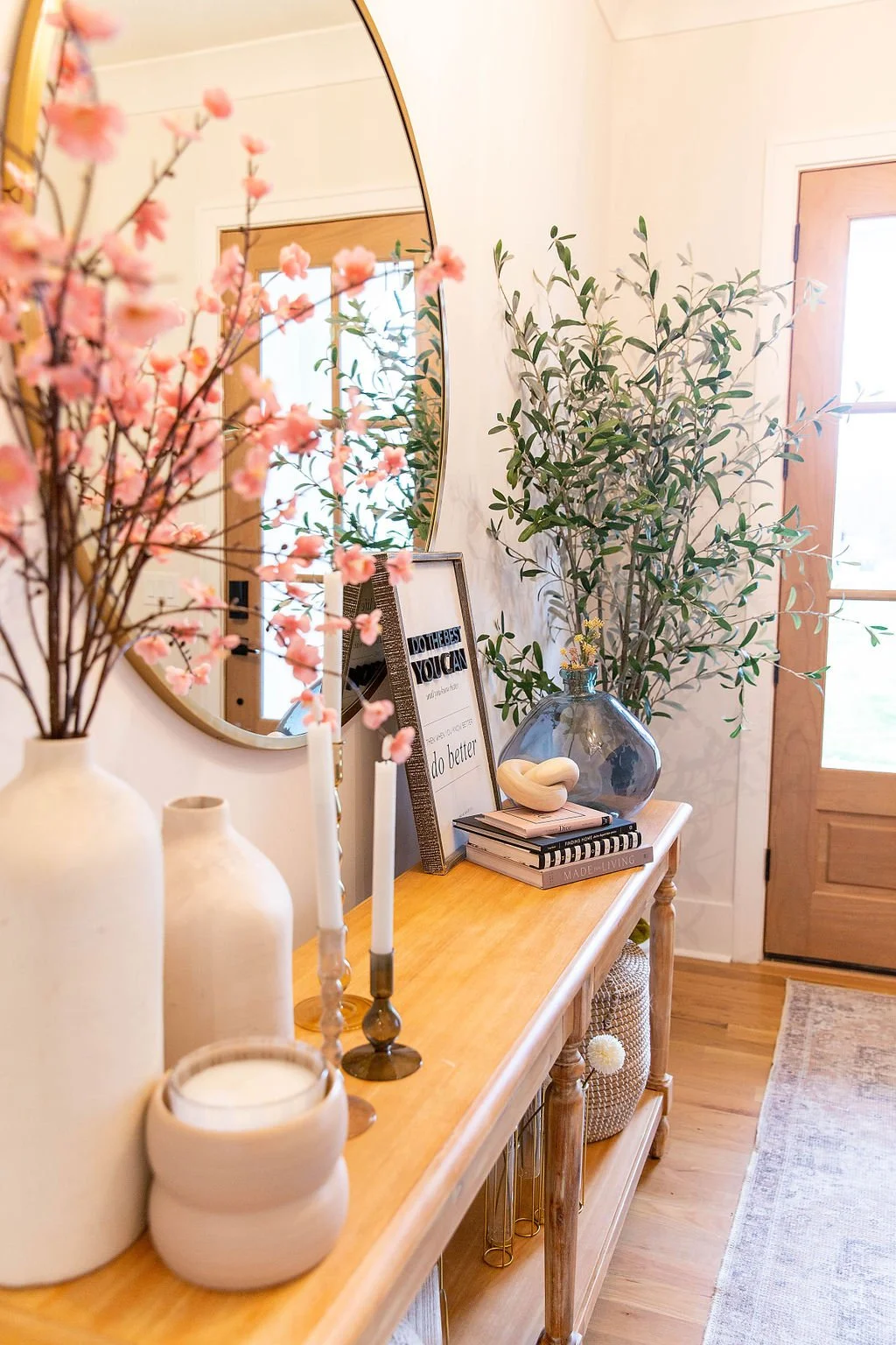 A wooden console table decorated with vases, books, candles, and framed artwork next to a mirror and a door, with a large leafy plant in a glass vase.