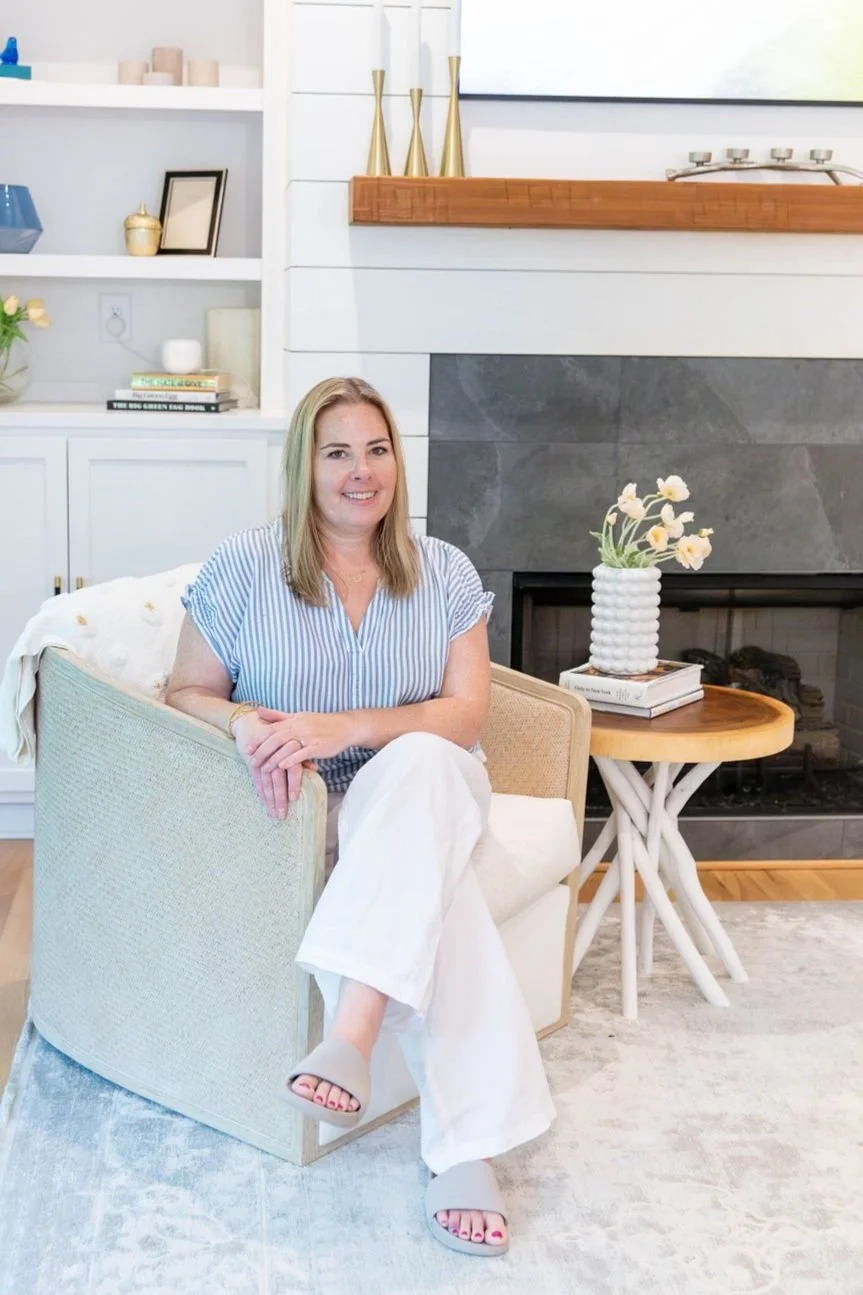 A woman with blonde hair wearing a blue and white striped top, white pants, and gray slippers is sitting on a beige chair in a living room with a modern fireplace, decorative objects, and bookshelf.