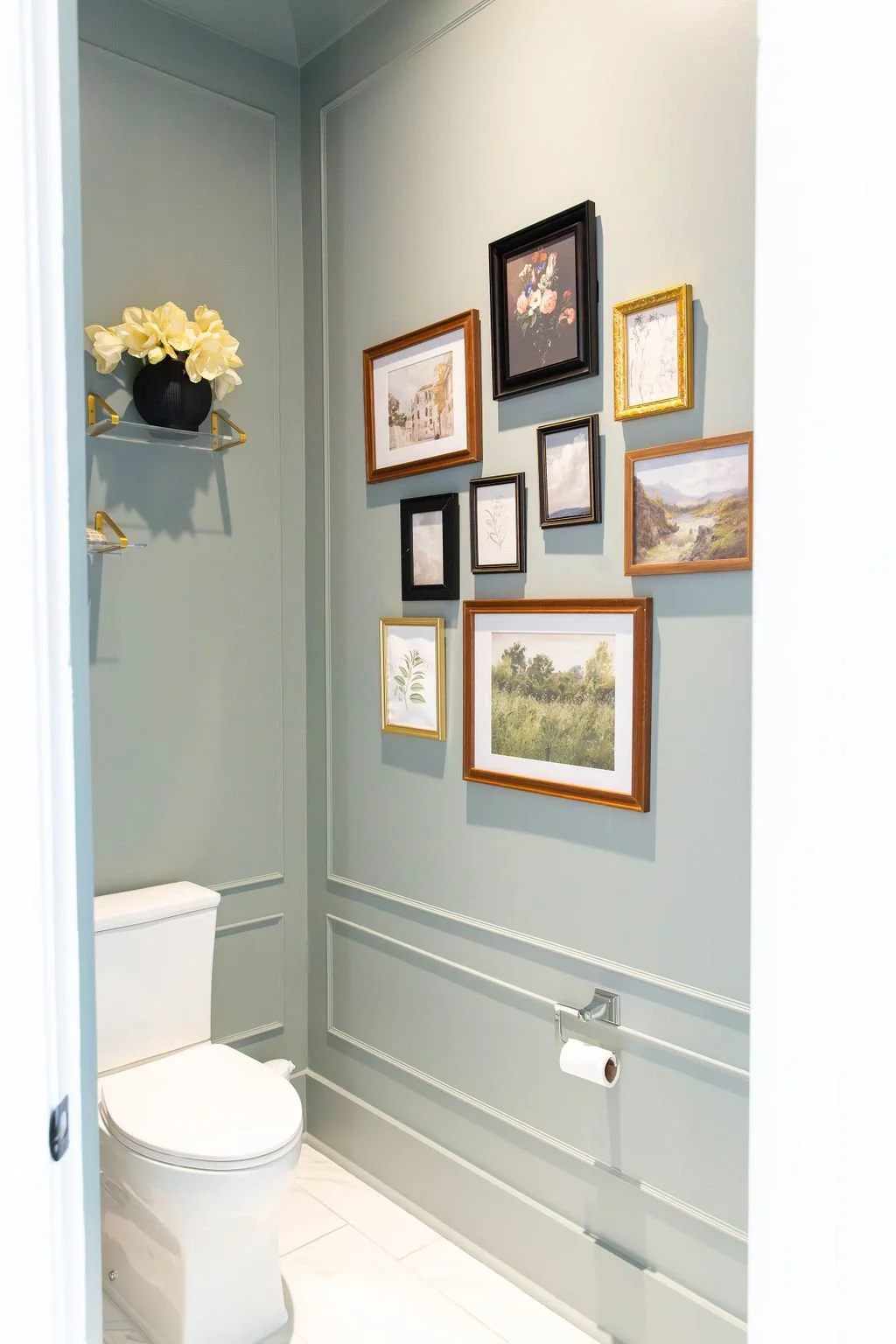 A bathroom with light green walls decorated with a gallery of various framed pictures, including landscapes, floral artwork, and sketches. A small shelf with a black vase holding cream-colored flowers is on the upper left corner. A white toilet with 
