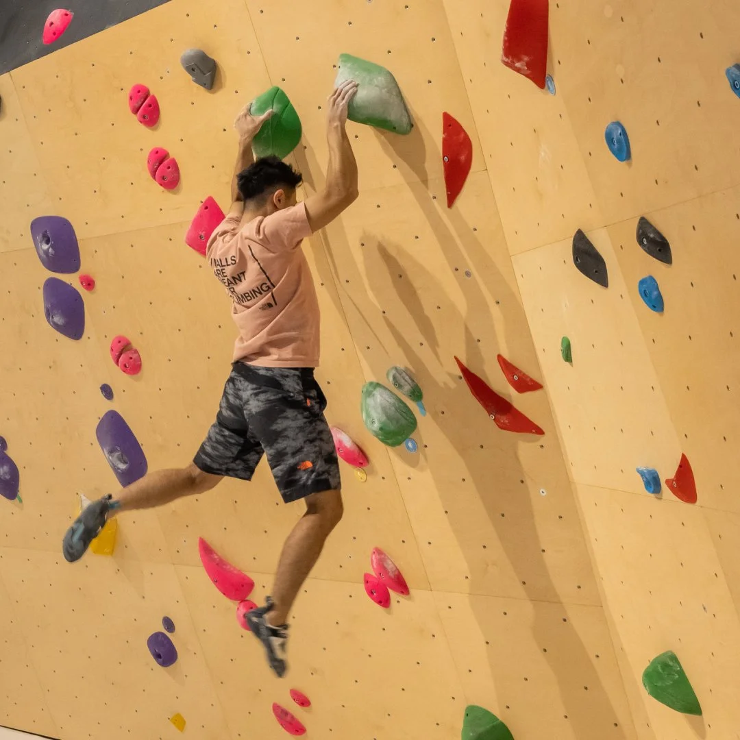 Young man climbing an indoor bouldering wall with colorful holds, including pink, green, red, purple, black, and blue.