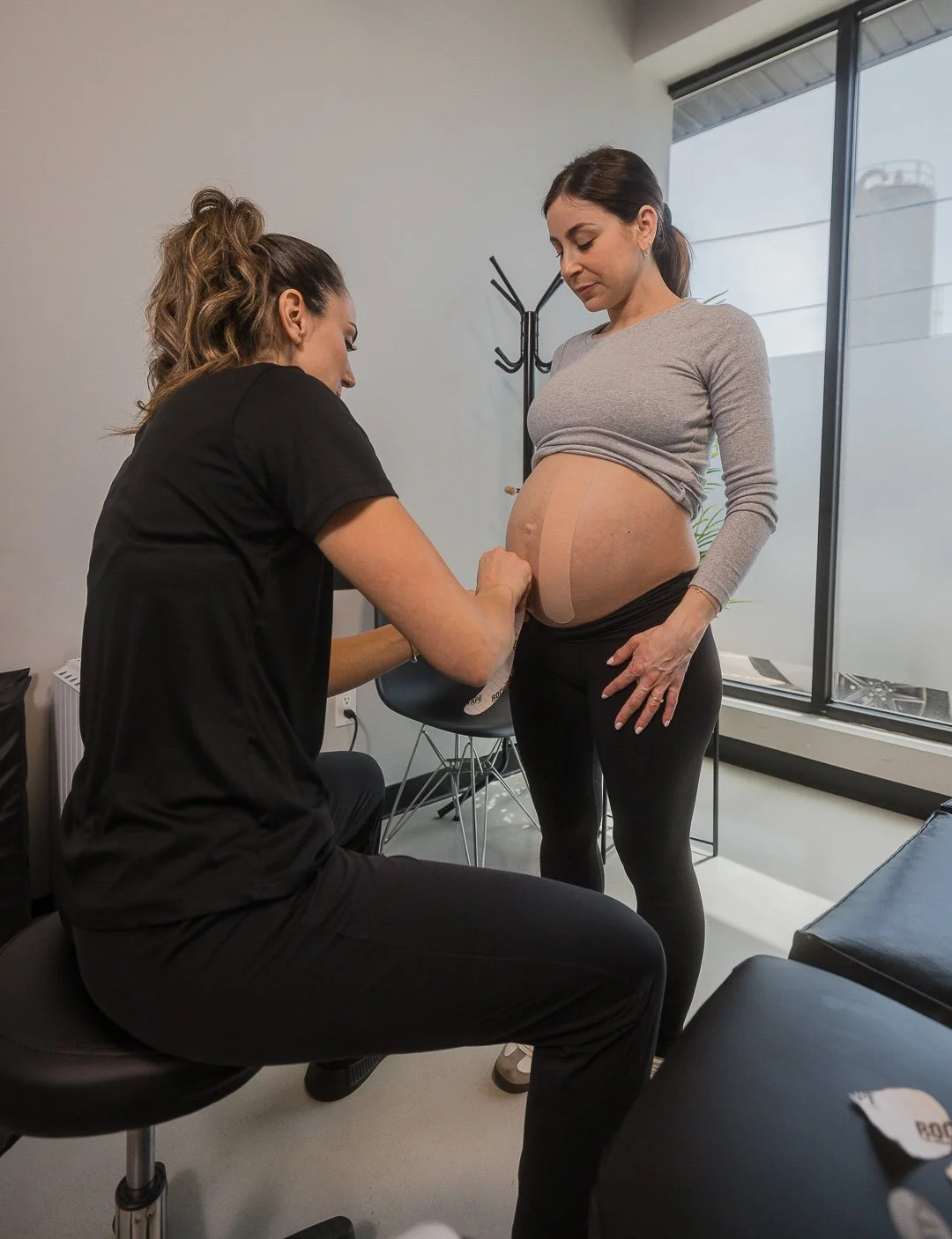 A pregnant woman standing while a healthcare professional applies KT tape to her abdomen inside a clinic.