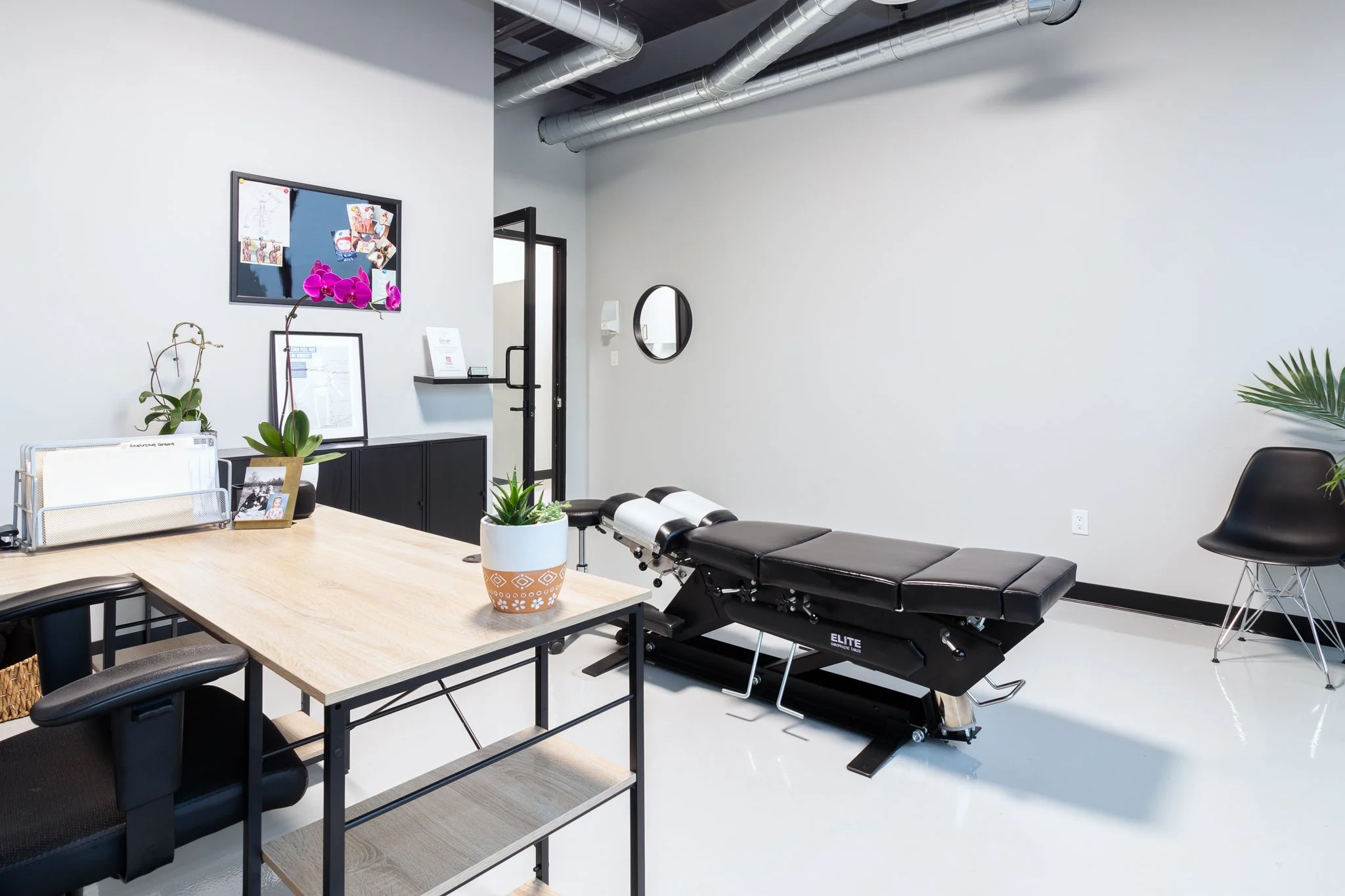A modern medical or chiropractic treatment room with a massage or adjustment table, a desk with black chairs, potted plants, framed photographs and artwork, a black cabinet, a round wall mirror, and a black chair, all in a bright space with white wal