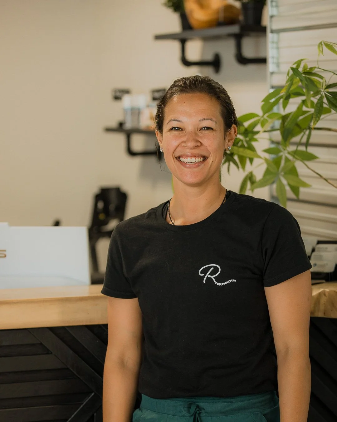 Smiling woman wearing a black T-shirt with a white 'R' and a small leaf design, standing in a modern indoor space with plants, shelves, and a wooden counter.
