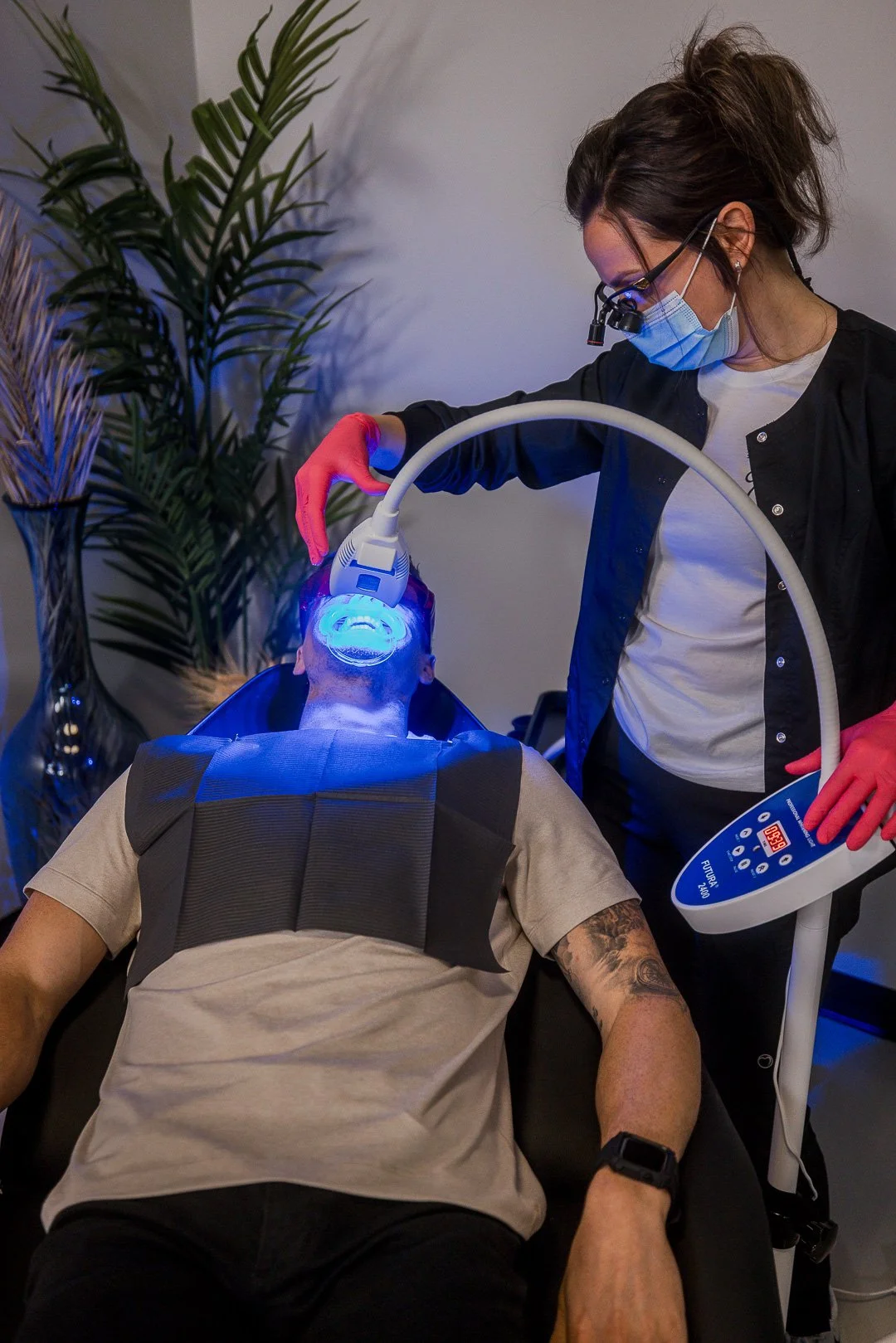 A woman receiving a dental treatment using a blue light on a patient who is lying down with a protective vest, while wearing a mask and glasses, in a dental clinic setting.