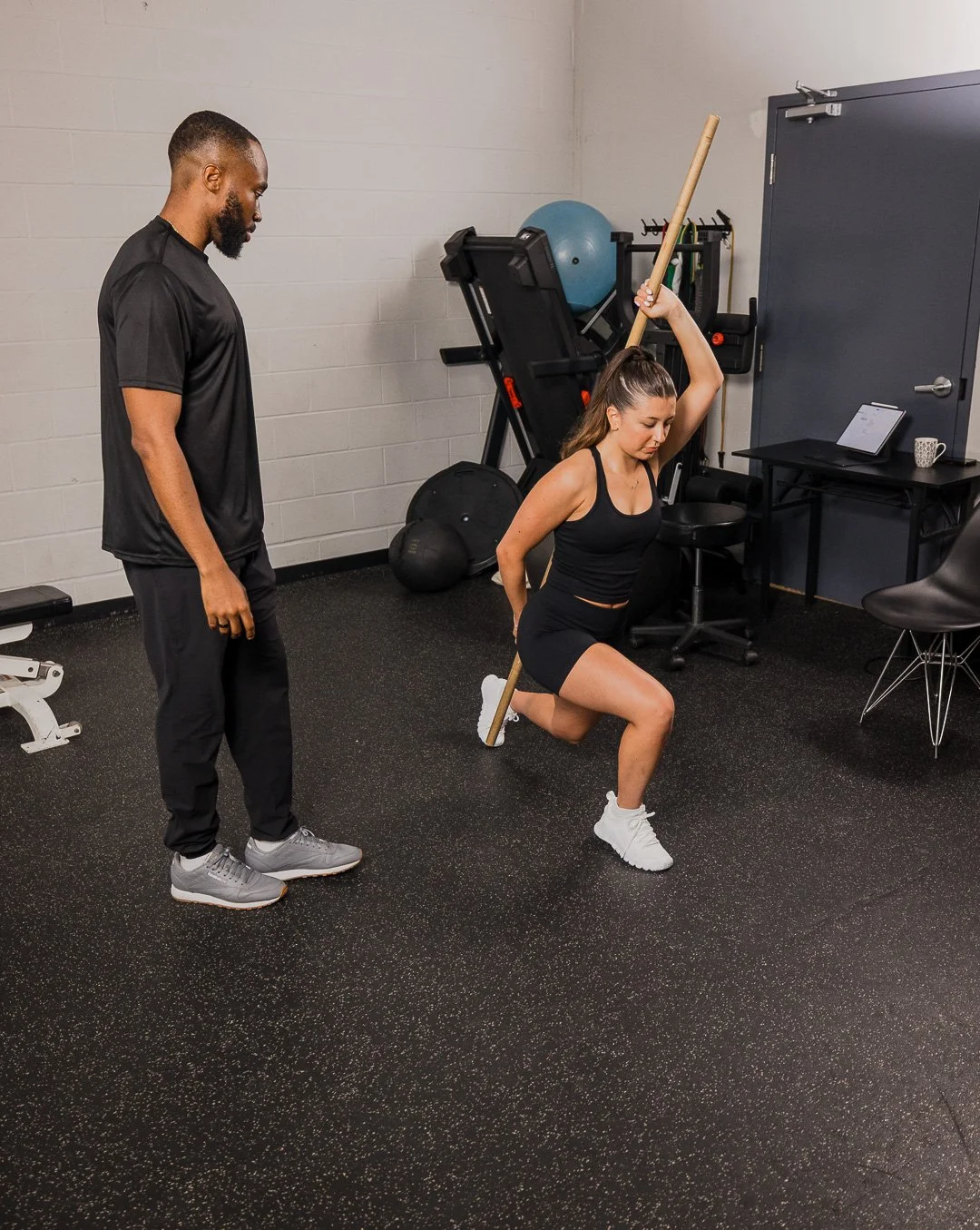 A woman in black workout clothes kneels on the ground holding a club overhead with one hand, under supervision by a man in black athletic attire in a gym setting.