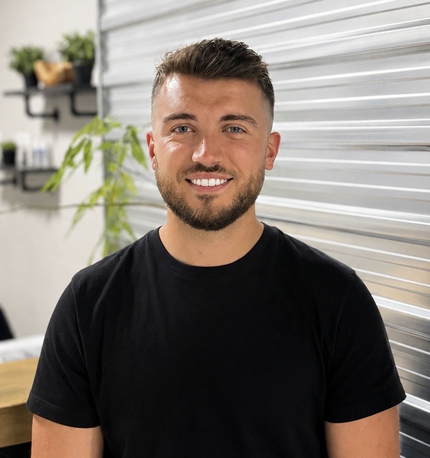 A smiling man with short brown hair and a beard, wearing a black t-shirt, standing indoors in front of a metallic wall with a green plant and shelves with small items in the background.