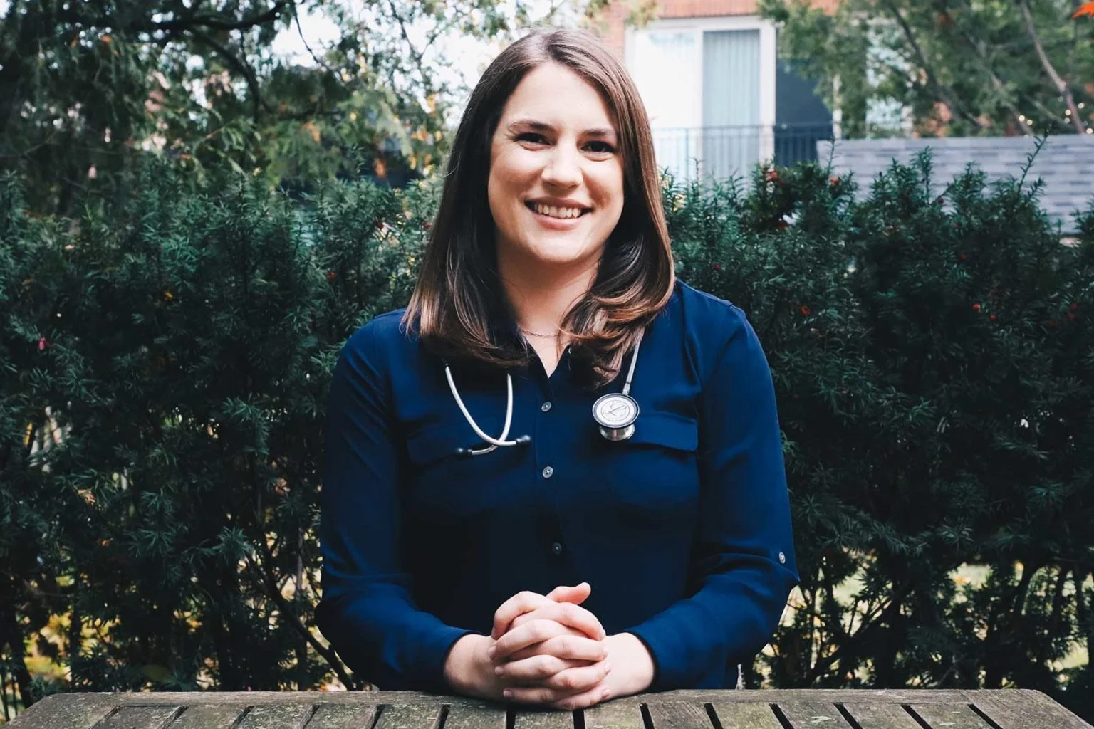 A woman with a stethoscope around her neck smiling outdoors in front of green bushes.