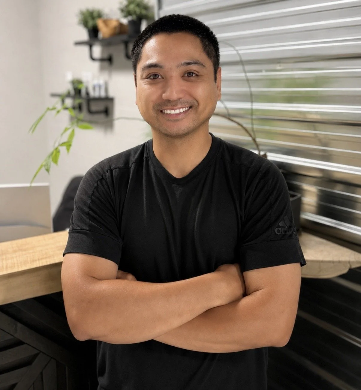 A smiling man with short black hair posing with arms crossed in a modern, well-lit room with plants and a window with horizontal blinds in the background.