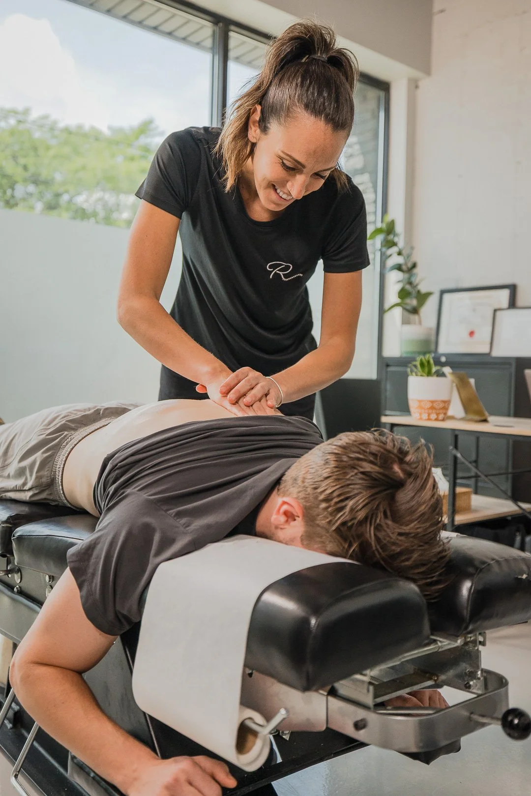 A woman giving a massage to a man lying face down on a massage table in a clinic or spa. The woman is smiling and appears to be focused on the massage.