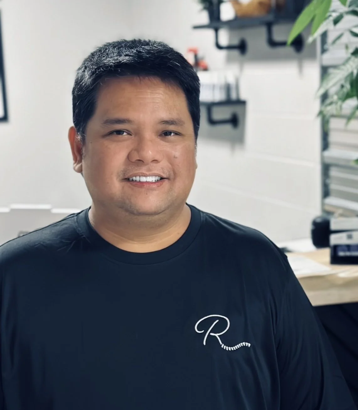 A smiling man with short black hair wearing a black t-shirt with white cursive letter 'R' sitting in a modern office with shelves, plants, and books in the background.