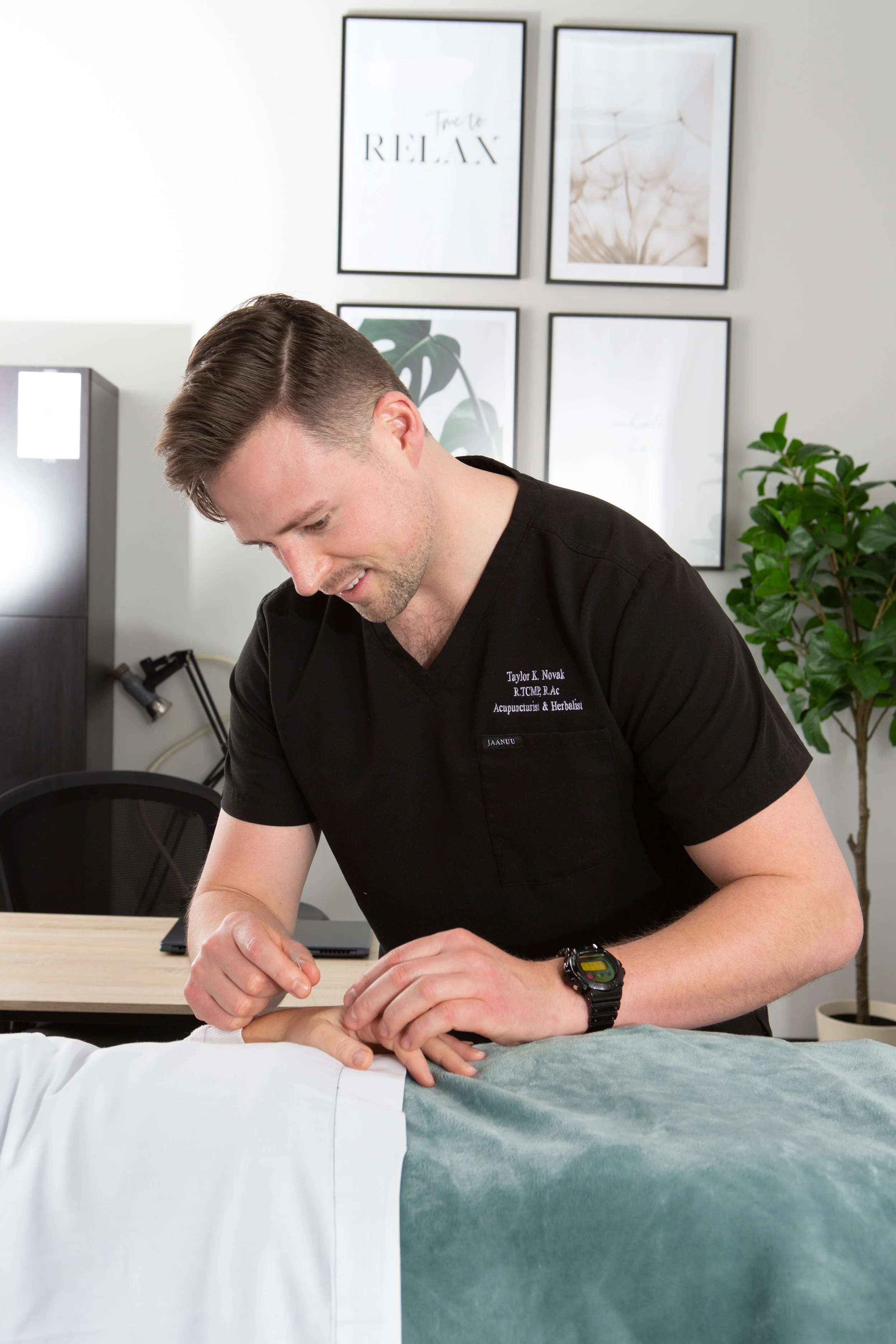A male therapist in black scrubs performs a manual therapy or massage on a patient's hand, who is lying on a table covered with a green sheet. The therapist has short brown hair, a digital watch, and a name tag on his scrubs. The room features framed artwork and a plant in the background.