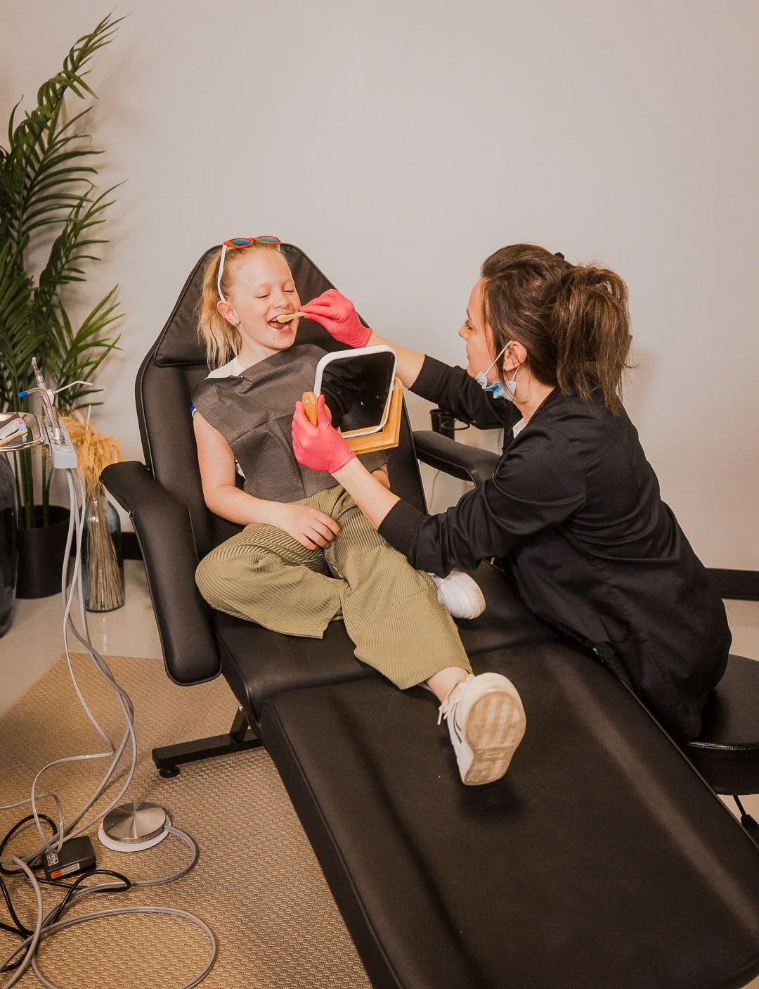 A young girl is lying back on a dental chair, smiling as a female dentist examines her mouth with a dental mirror. The dentist is wearing a face mask and pink gloves, and the girl is holding onto her lap. There are dental equipment and cords on the floor, and a large plant in the background.