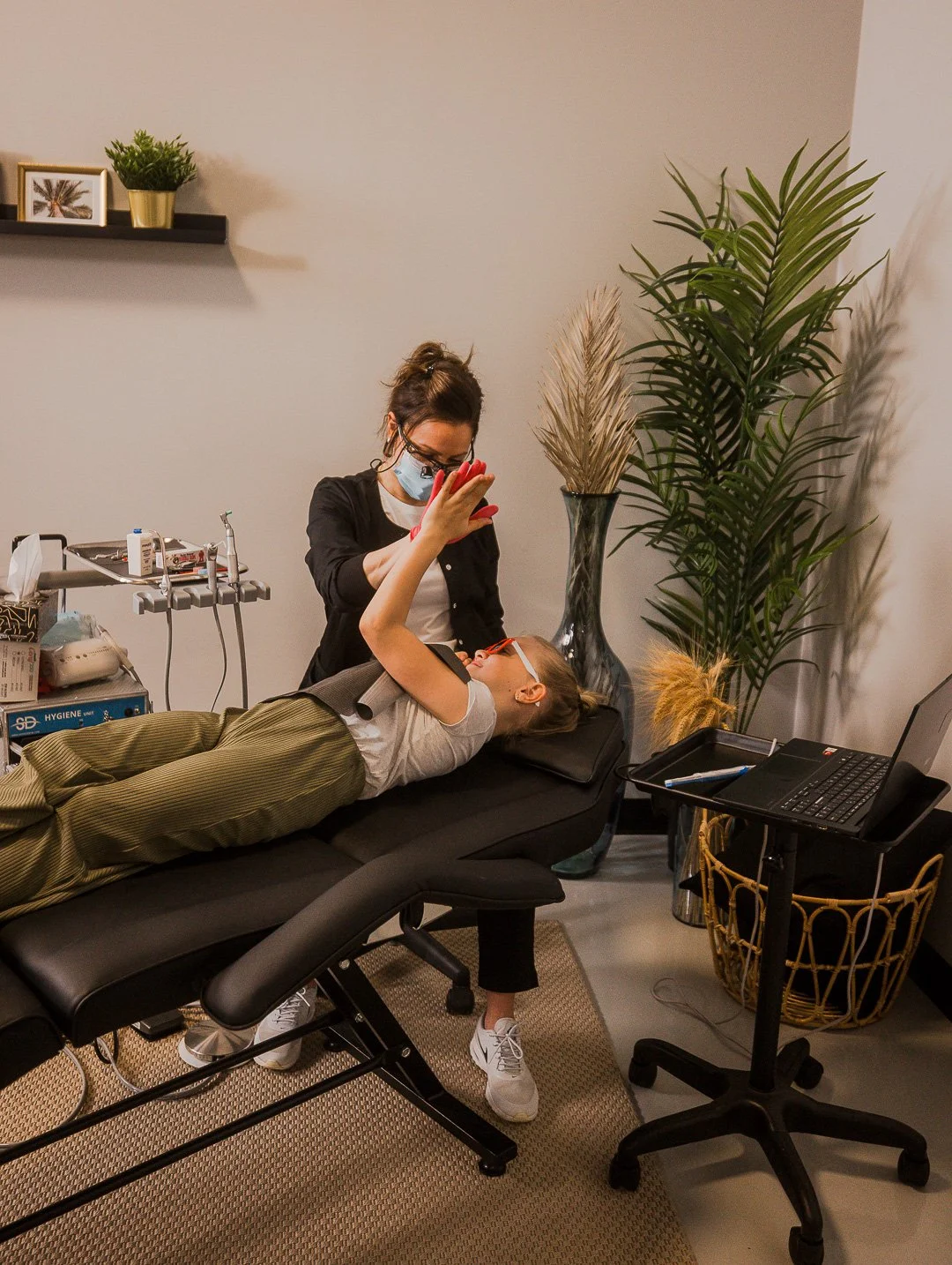 A woman lying on a medical exam table, wearing glasses and a gray shirt, taking a selfie with a smiling woman standing next to her, who is wearing a face mask and black outfit. There are large potted plants, a laptop, and medical equipment in the room.