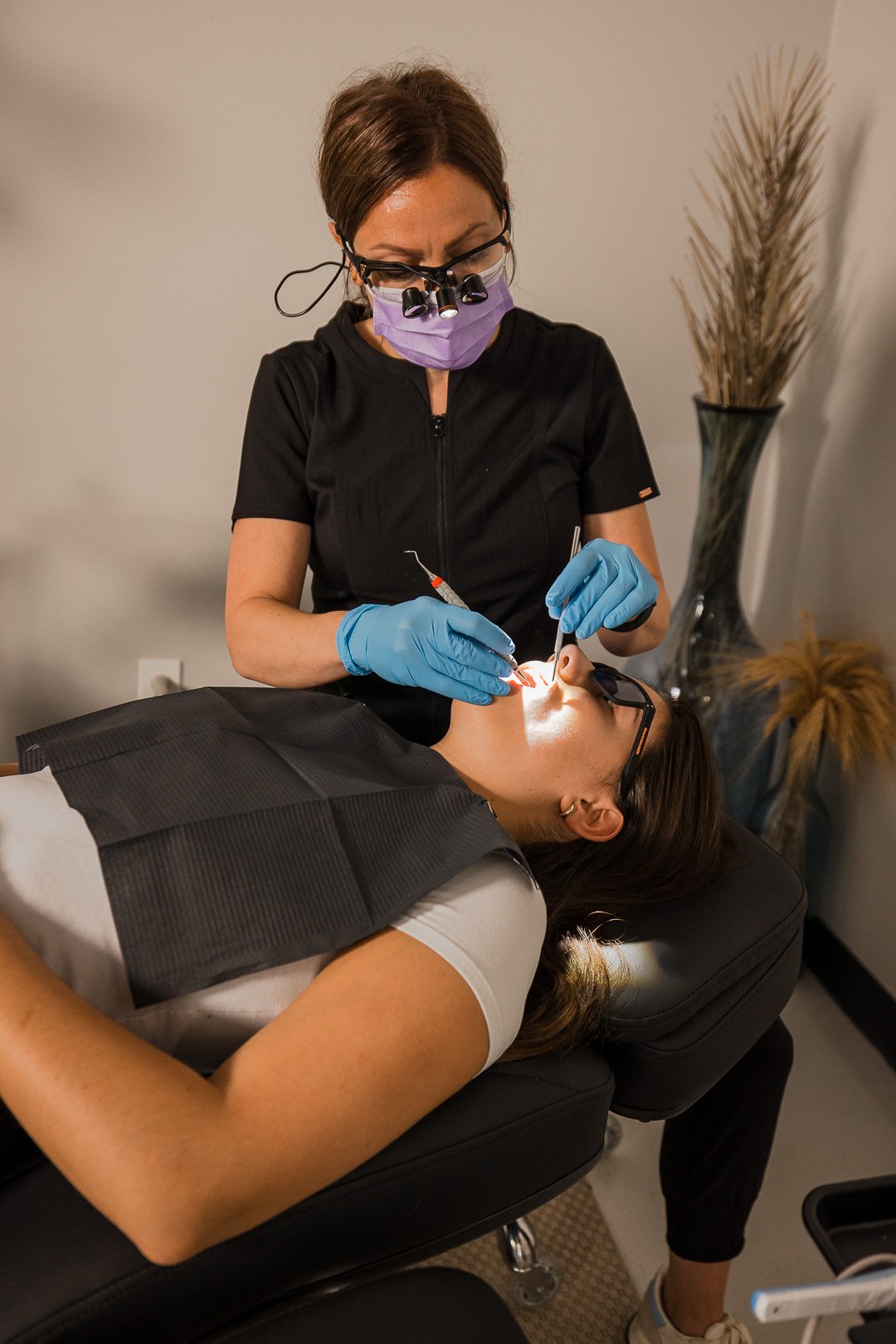 A female dentist wearing magnifying glasses and a purple mask performs a dental procedure on a female patient lying in a dental chair, with a lamp illuminating her mouth.