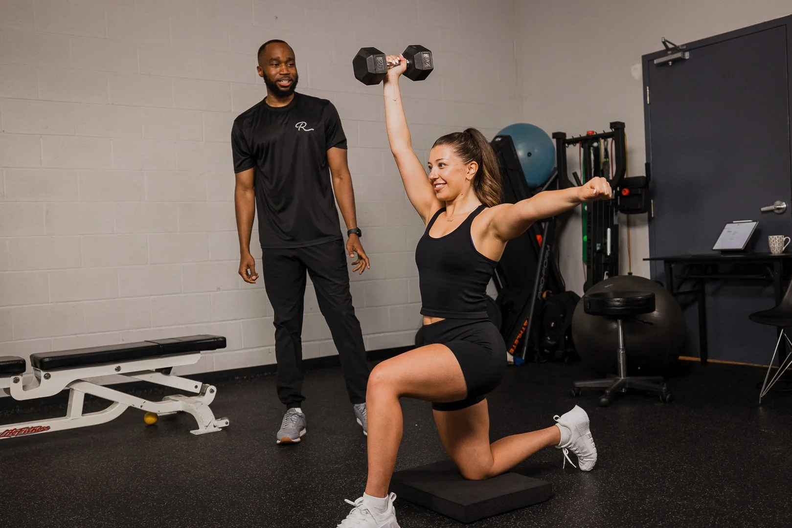 A woman is kneeling on one knee on a exercise mat, holding a dumbbell overhead in a lunge position. A man is standing nearby, observing and smiling in a gym with various equipment and a gray brick wall.