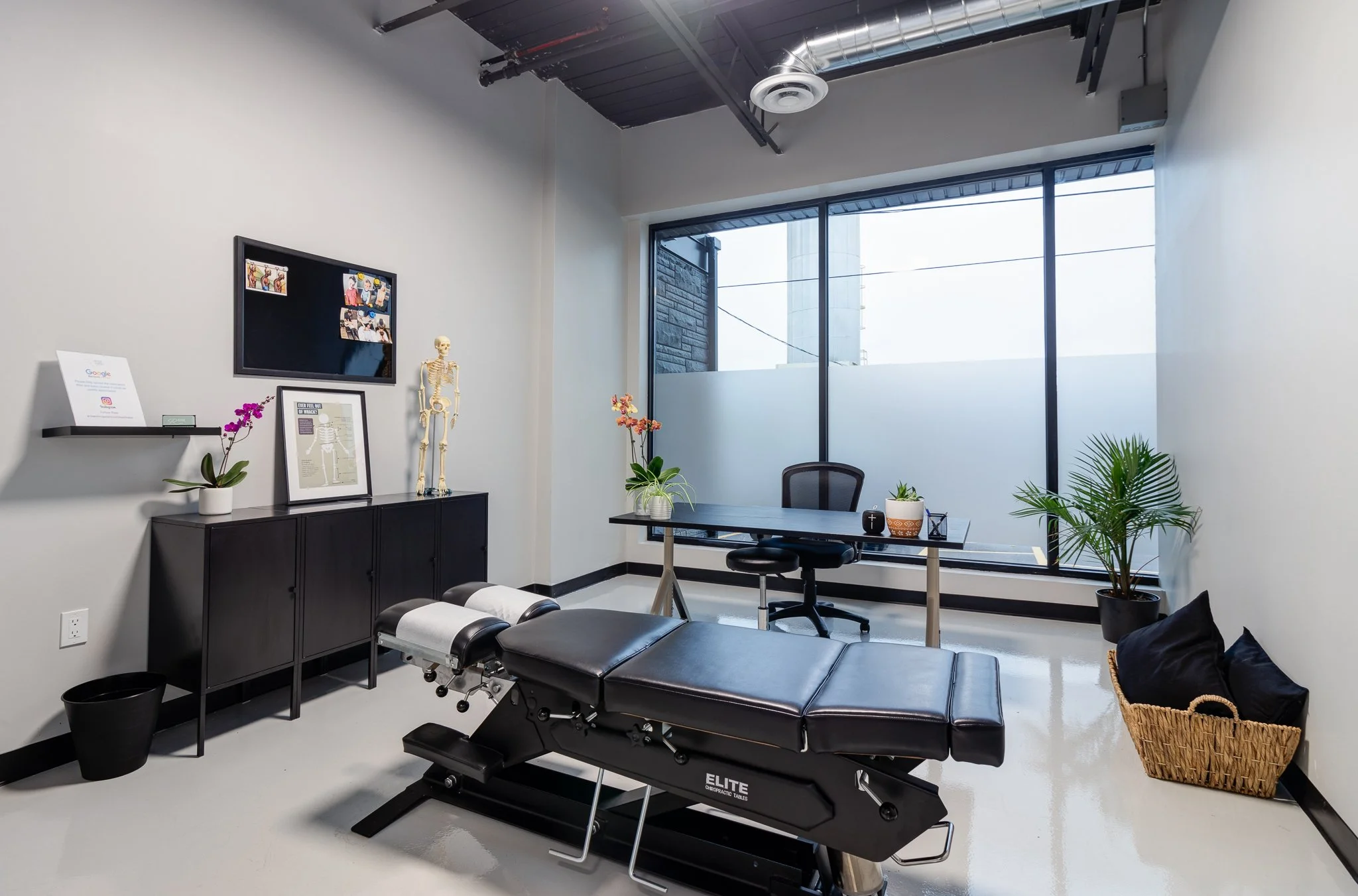 Modern chiropractor's office with treatment table, black cabinet, desk with plants, large window, and medical decor.