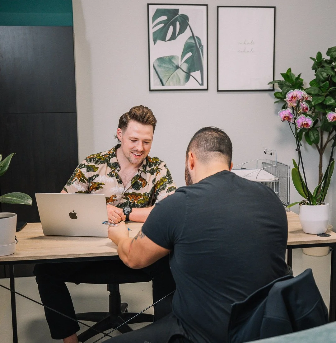 Two men sitting at a desk having a discussion in an office with plants and framed leaf artwork on the wall.