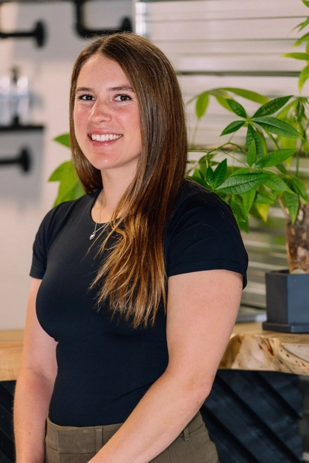 A woman with long brown hair smiling, wearing a black t-shirt and khaki pants, standing next to a green plant in an indoor setting.