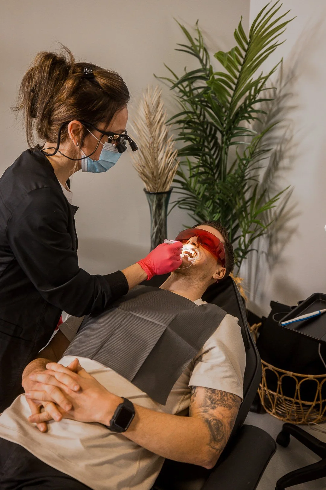Dentist wearing mask and gloves performs dental work on patient in red protective glasses, lying back in dental chair.