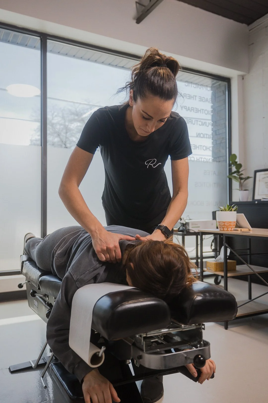 A chiropractor adjusting a woman's neck on a chiropractic table in an office with large windows, desks, and potted plants.