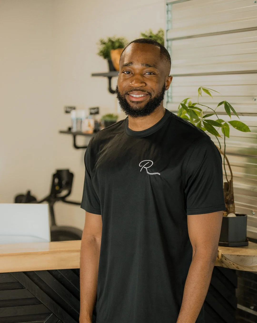 A smiling man with a beard in a black t-shirt standing indoors near plants and the counter.