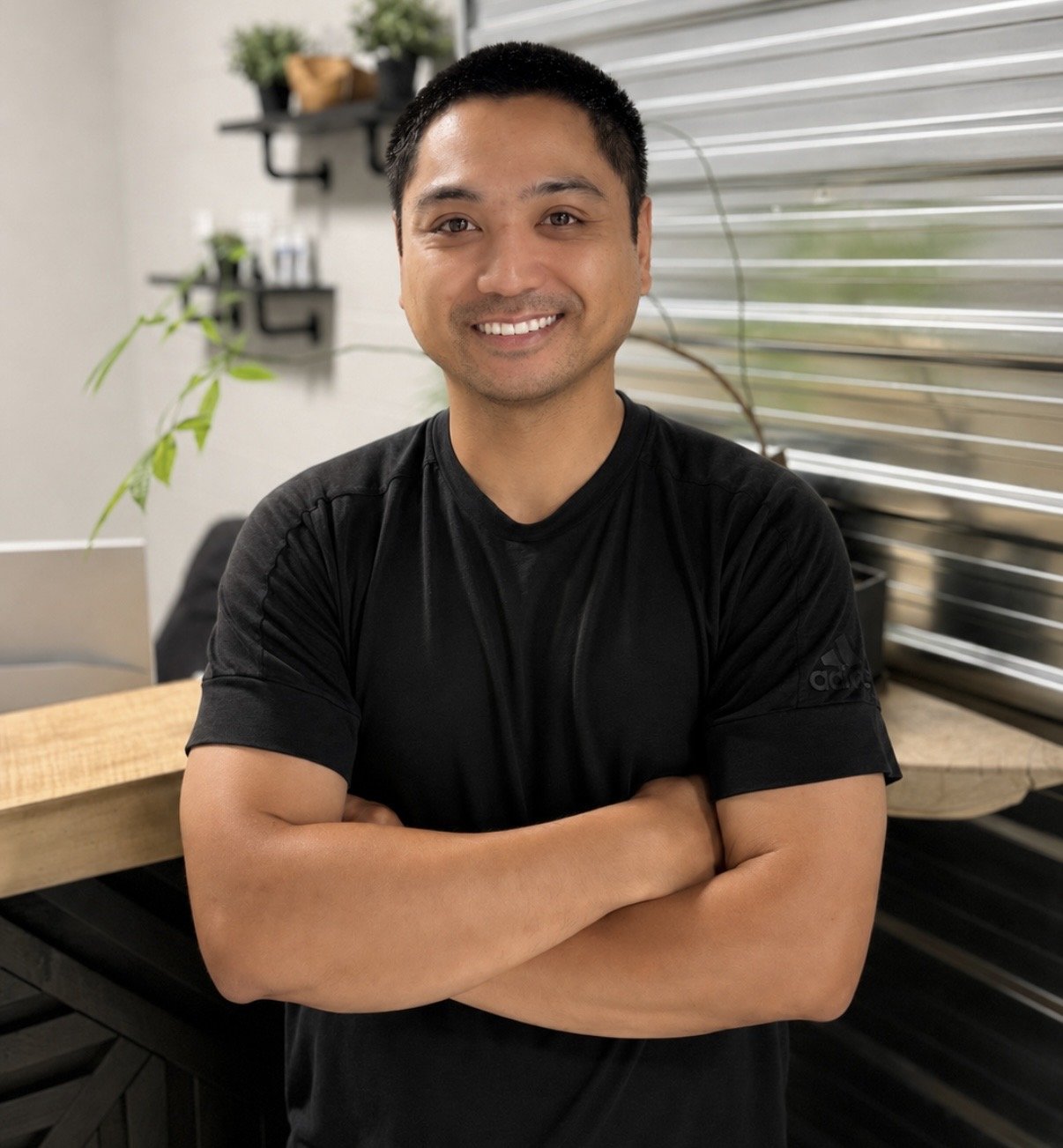 A man with short black hair smiling with arms crossed, standing in a modern indoor space with plants and a window with blinds in the background.