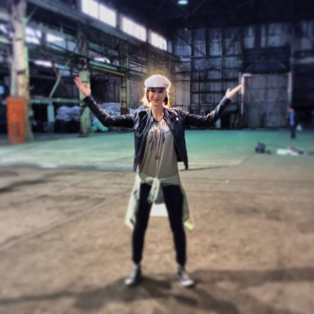 Person in a leather jacket and cap standing with arms outstretched in a large industrial warehouse.