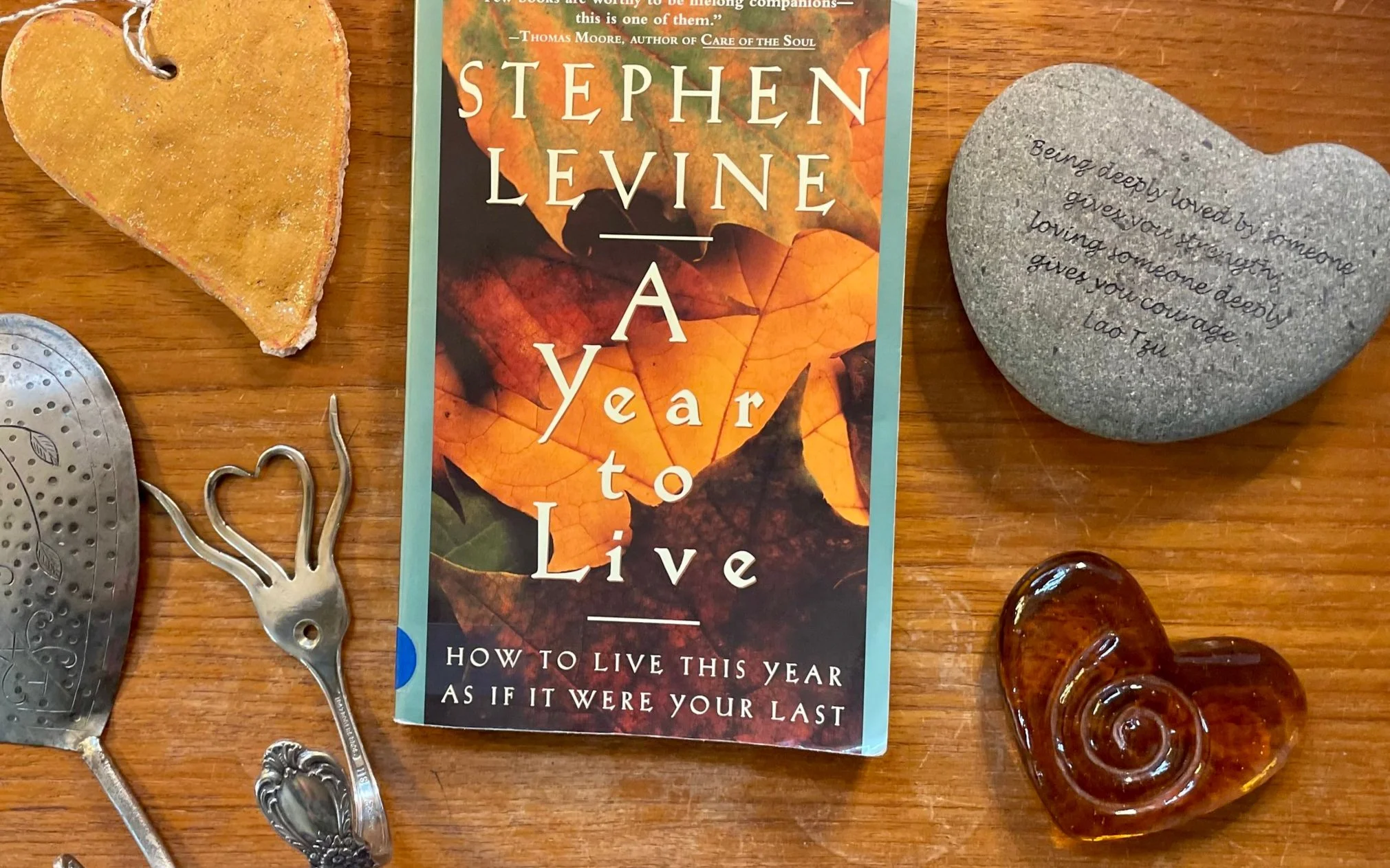 Image of a book titled 'A Year to Live' by Stephen Levine placed on a wooden surface. Surrounding the book are various objects including a ceramic heart with an inspiring quote, a pie slice, a metal decorative item, a small glass bottle, and a few other miscellaneous objects.