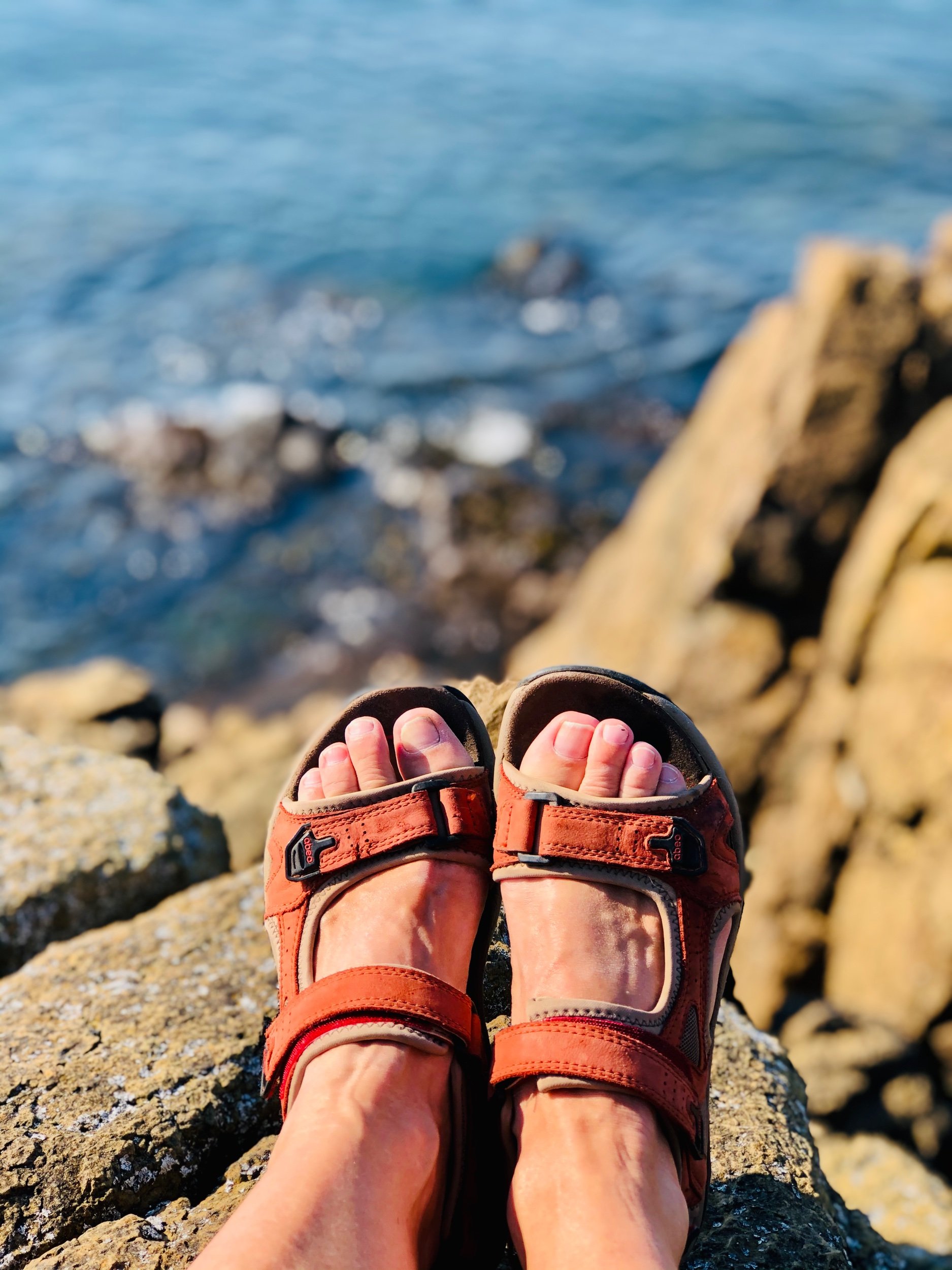 Person wearing red hiking sandals with toes visible, sitting on rocks by the water, with the sea and blurred rocks in the background.