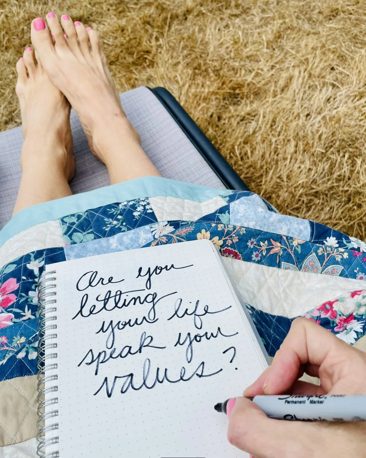 A person sitting on a lounge chair outdoors with bare legs and painted toenails, holding a notebook and writing with a black marker. The notebook has a handwritten message that reads, 'Are you letting your life speak your values?'. The person is wear